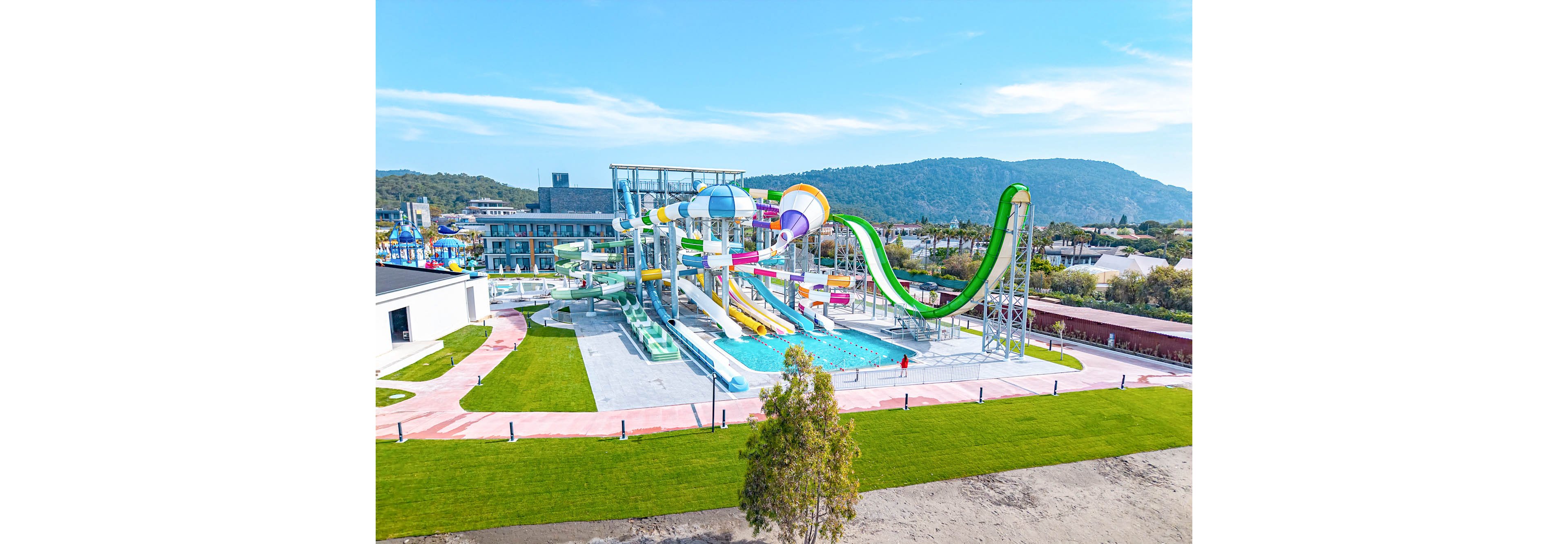 A large outdoor water park features multiple colorful water slides converging into a central pool. The park is surrounded by neatly maintained green lawns and walking paths. In the background, low-rise resort buildings and forested hills create a scenic backdrop under a clear blue sky. The bright colors and sunny weather give the scene a fun, family-friendly atmosphere.