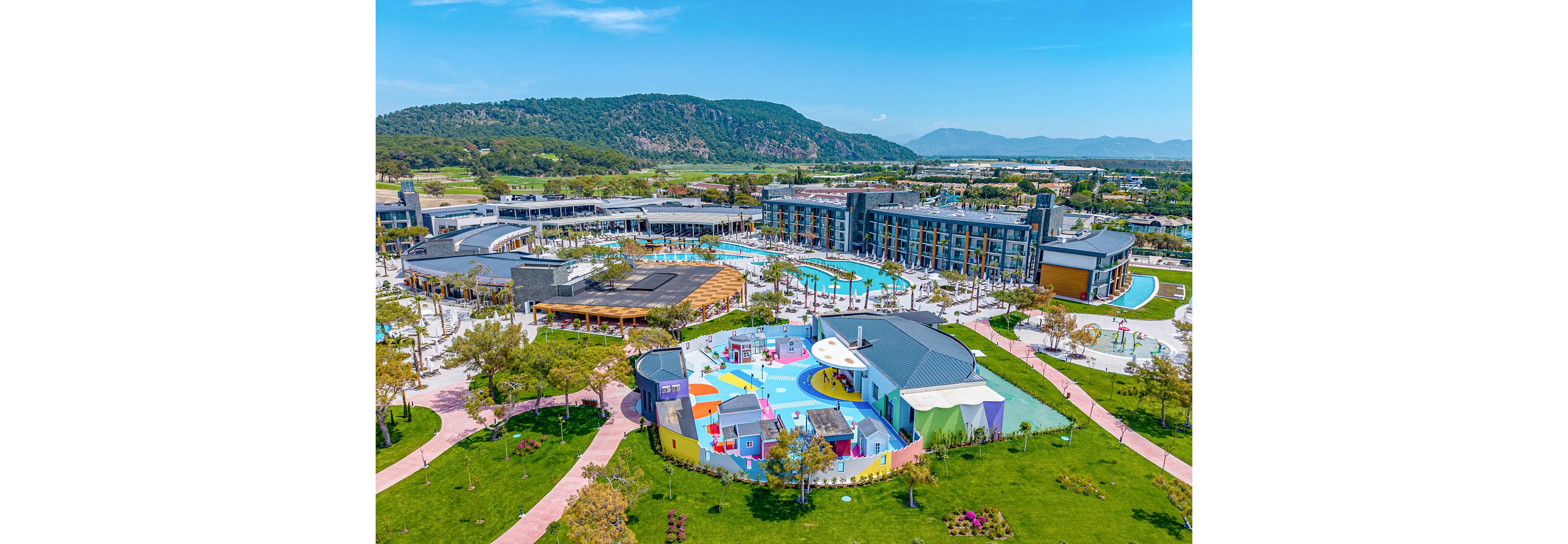 An aerial view showcases a large modern resort complex with multiple buildings and outdoor swimming pools. In the foreground, a colorful children’s play area with bright structures and slides stands out. Curved walking paths, lawns, and young trees surround the resort, creating a landscaped park atmosphere. Mountains and open countryside form a scenic natural backdrop under a clear blue sky.