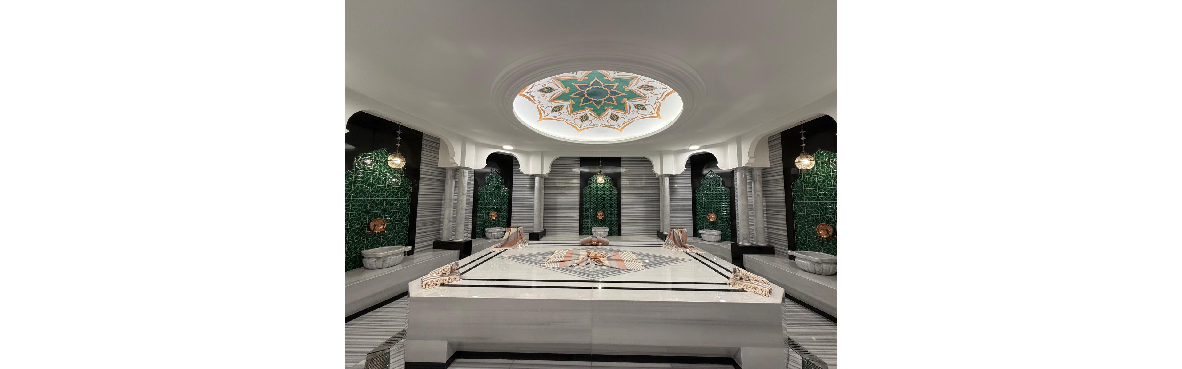 The image showcases an opulent Turkish hammam featuring a large central marble platform surrounded by intricately tiled green alcoves and elegant marble basins. The ceiling displays a striking geometric pattern in green, gold, and white, adding to the luxurious ambiance. Hanging lamps and arched columns enhance the traditional and serene atmosphere. The space is empty, emphasizing the architectural details and symmetry.