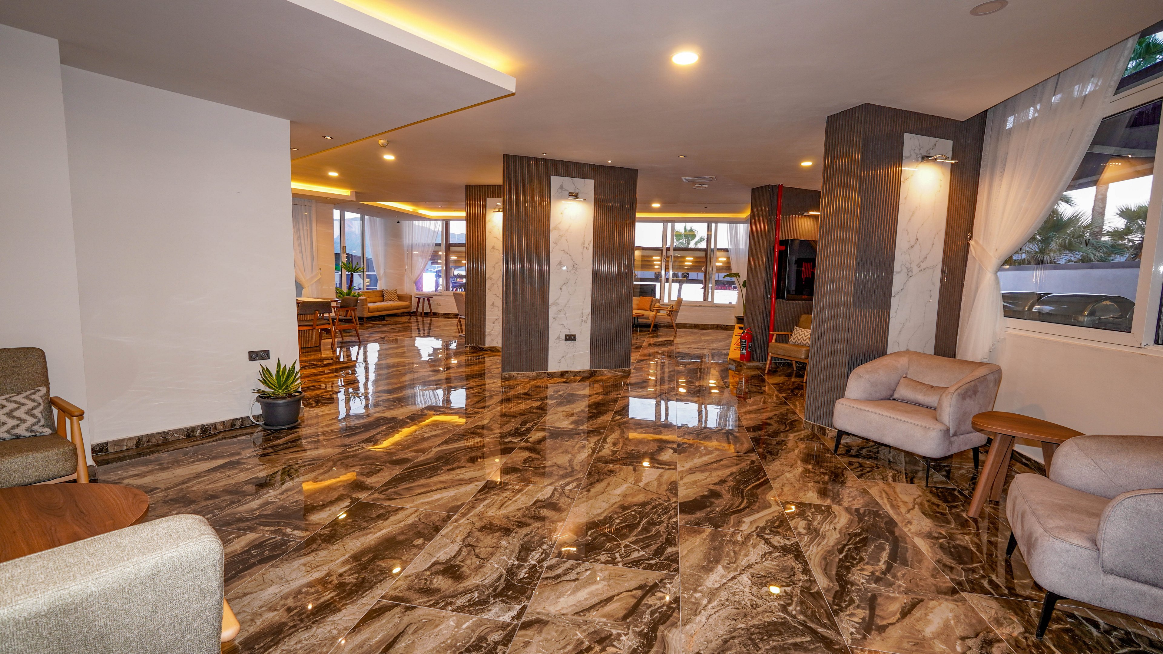 Spacious contemporary lobby interior featuring a highly polished marble floor with bold veining. Neutral-toned armchairs and small tables are arranged along the perimeter, creating relaxed seating areas. Large windows and recessed ceiling lights provide warm, even illumination throughout the space. The scene suggests a modern hotel or apartment building reception area in the evening.