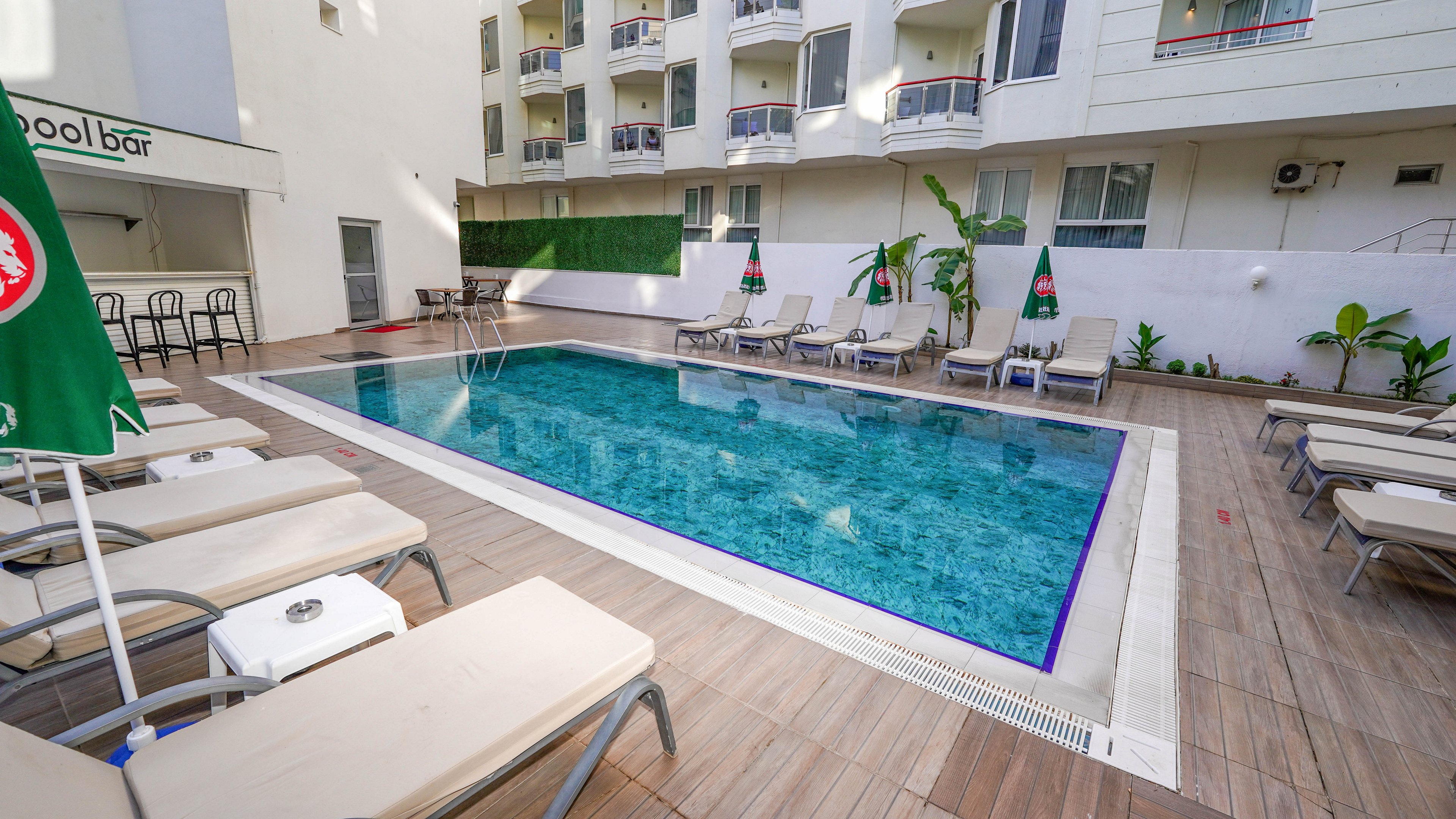 An outdoor rectangular swimming pool is surrounded by cushioned lounge chairs on a wooden deck at a modern hotel. The pool area is bordered by a white multi-story building with balconies overlooking the water. Green umbrellas and a small bar area add to the leisure setting. The scene is bright and clean, suggesting a relaxing vacation or business-stay environment.