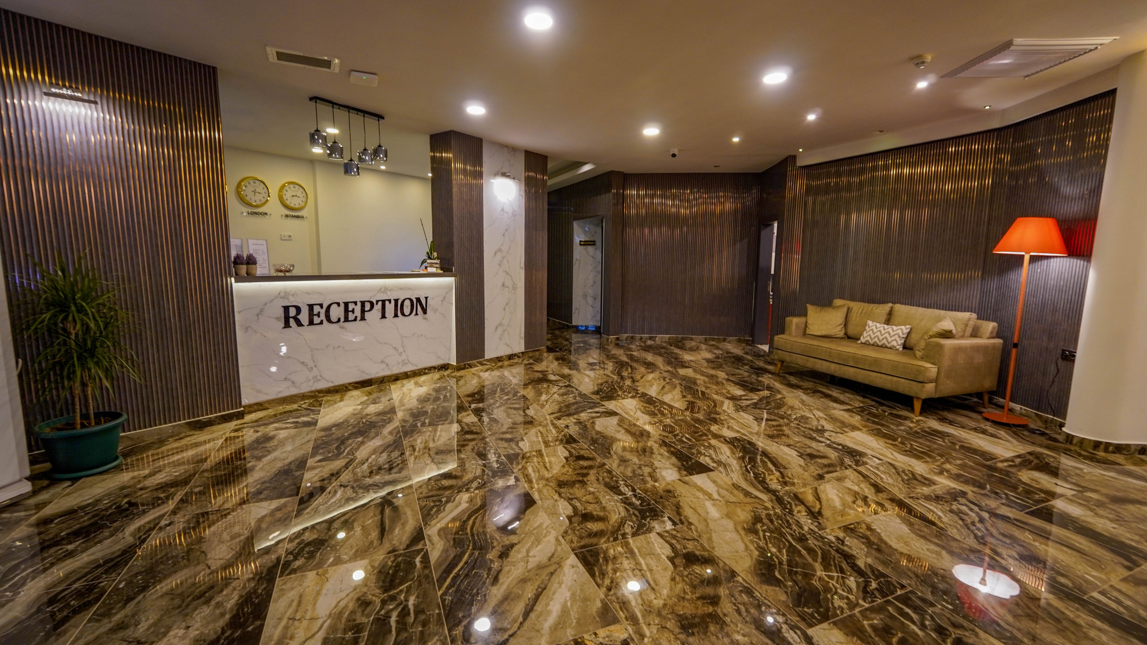 A contemporary hotel reception desk stands at the back of a spacious lobby. The area features a glossy marble floor with rich brown and beige patterns. Warm lighting, a beige sofa with cushions, and a red floor lamp create a welcoming atmosphere. Vertical wall panels and decorative clocks add to the modern interior design.