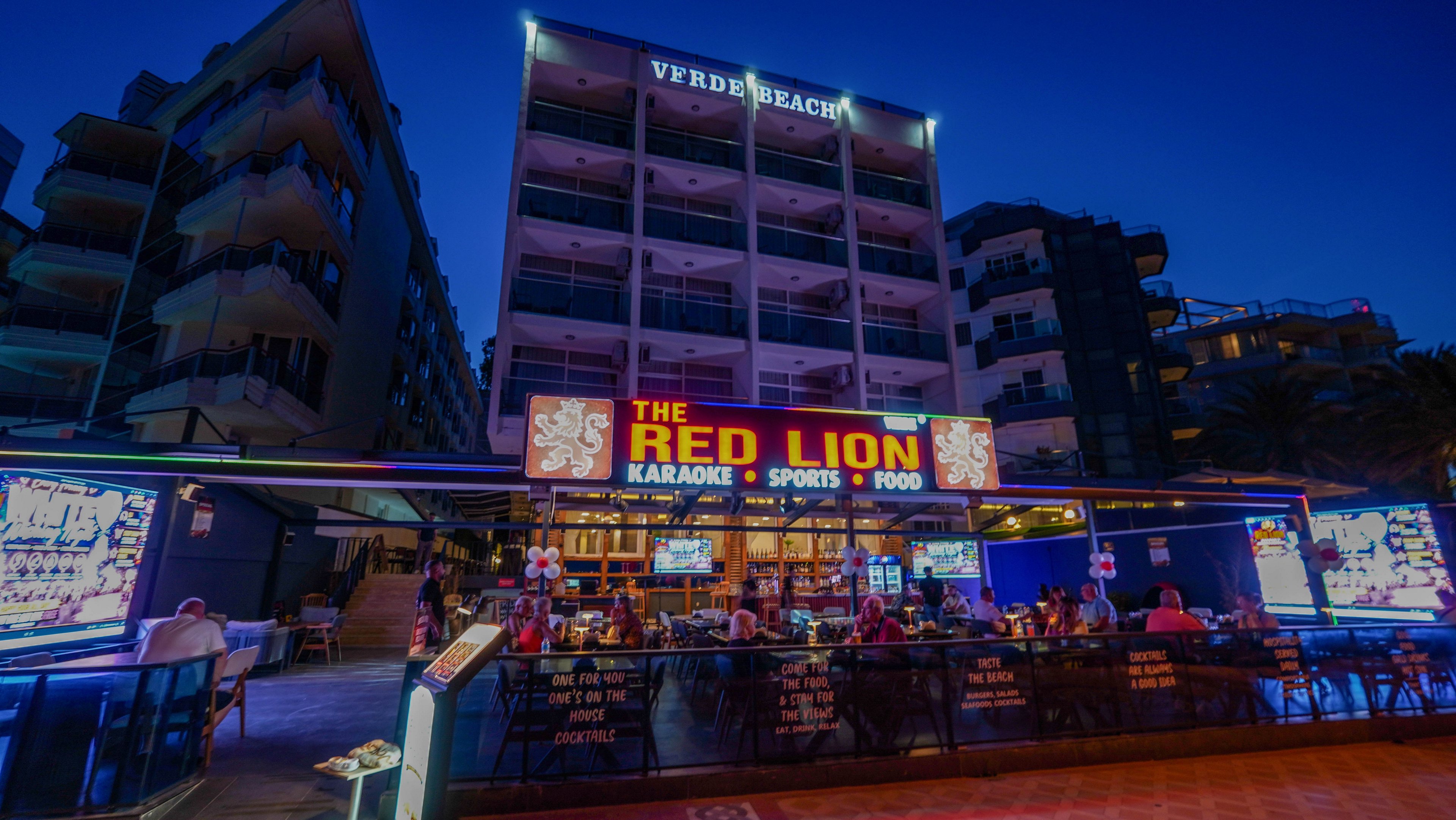 A lively bar called The Red Lion is illuminated at night beneath a tall hotel building labeled Verde Beach. Patrons sit at outdoor tables enjoying drinks and food under bright neon signage. The bar advertises karaoke, sports, and food in bold yellow and blue text. Surrounding apartment and hotel buildings frame the scene against a deep blue evening sky.