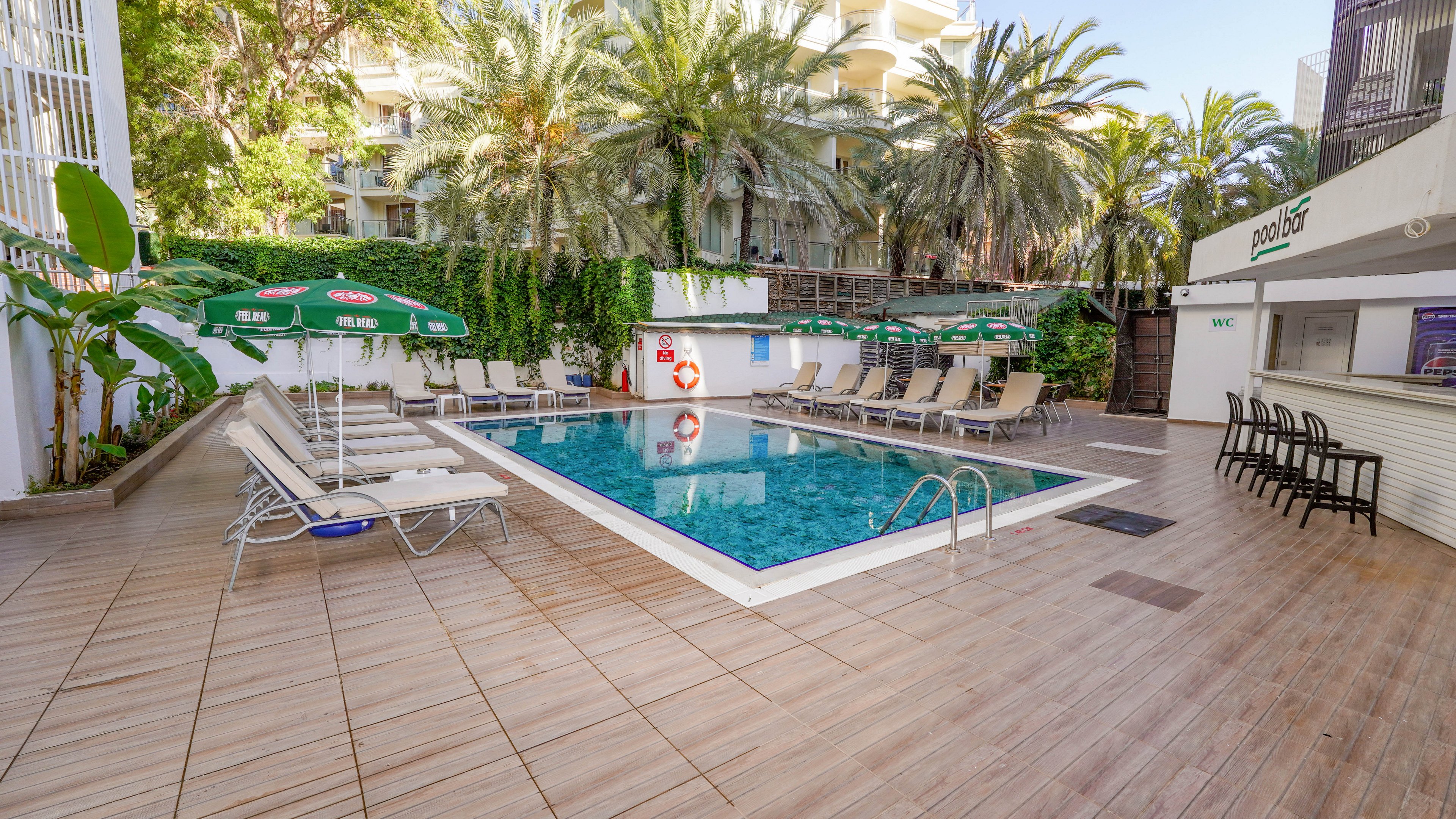 Outdoor hotel swimming pool surrounded by sun loungers and green umbrellas on a tiled terrace. A small pool bar stands to the right with high stools and a shaded counter. Lush palm trees and greenery create a tropical atmosphere around the courtyard. The scene is quiet and empty, suggesting a relaxing vacation setting.