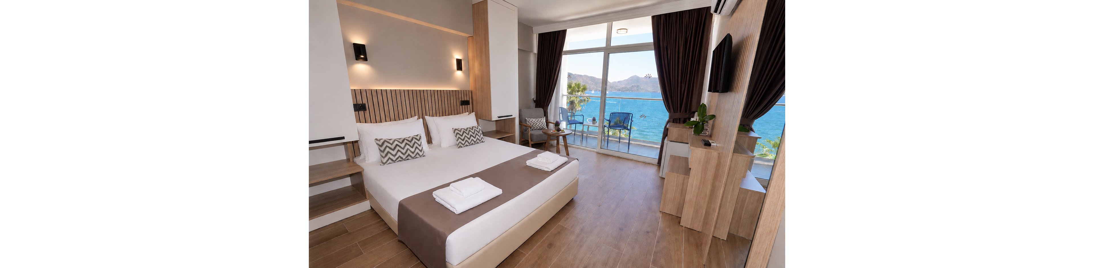 A contemporary hotel bedroom features a large double bed with crisp white linens and folded towels. Warm wood accents, minimalist furniture, and soft lighting create a clean, inviting atmosphere. Floor-to-ceiling windows open onto a balcony with chairs overlooking a bright blue ocean and distant mountains. Dark curtains frame the view, while a wall-mounted television and small desk complete the modern interior.
