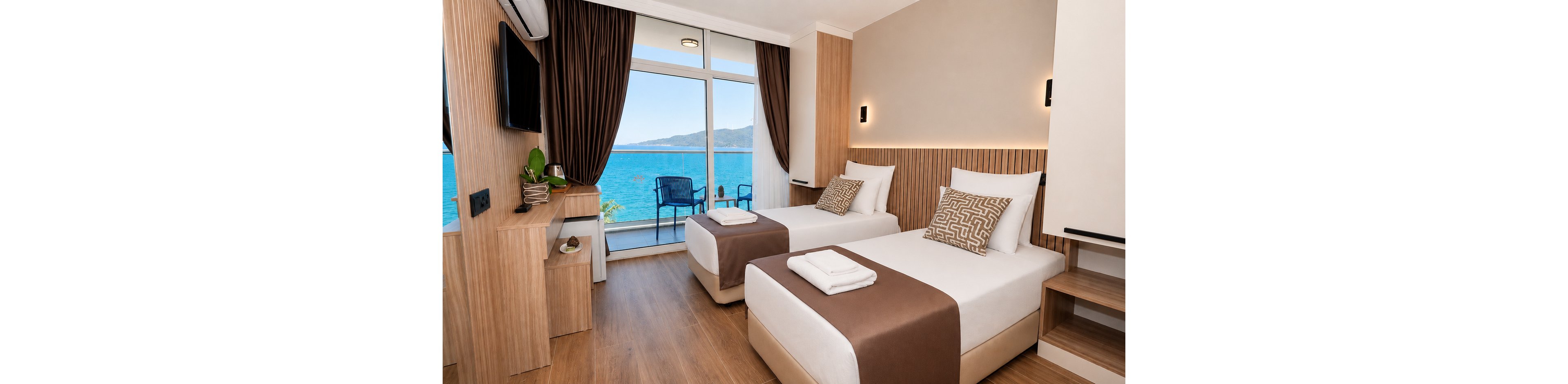 Contemporary hotel room featuring two neatly made twin beds with brown runners and patterned cushions. The space includes light wood furniture, a wall-mounted television, and a small desk with decor. Large glass doors open to a balcony with blue chairs overlooking a calm ocean and distant mountains. Warm lighting and neutral tones create a clean, relaxing atmosphere ideal for vacation or business travel.