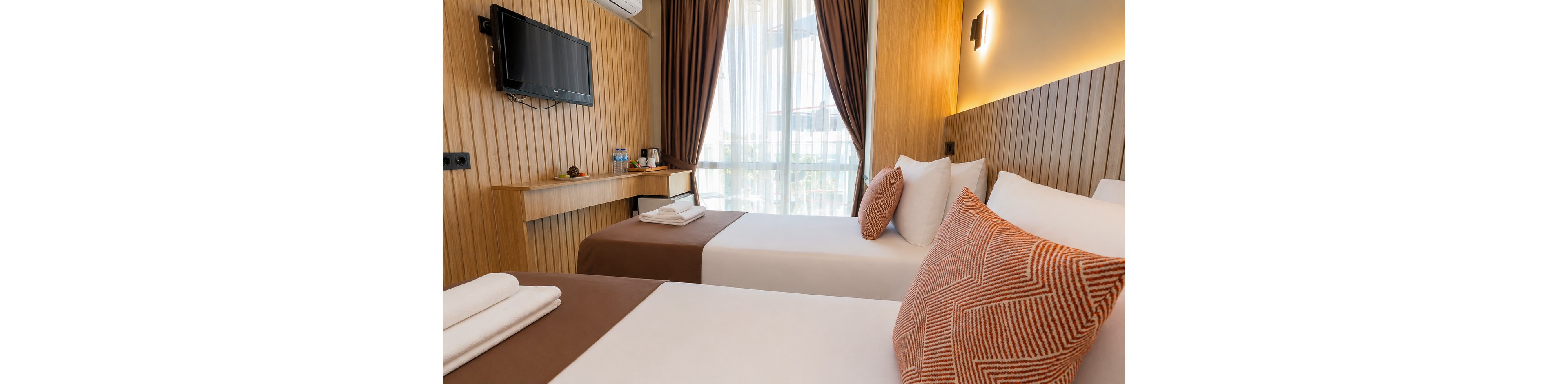 Contemporary twin beds are neatly arranged in a compact hotel room with warm wooden paneling. Large windows with sheer curtains and dark drapes allow natural light to brighten the space. A wall-mounted television, small desk, and complimentary beverage tray add functional comfort. Clean white linens, folded towels, and patterned accent pillows create a welcoming and cozy atmosphere.