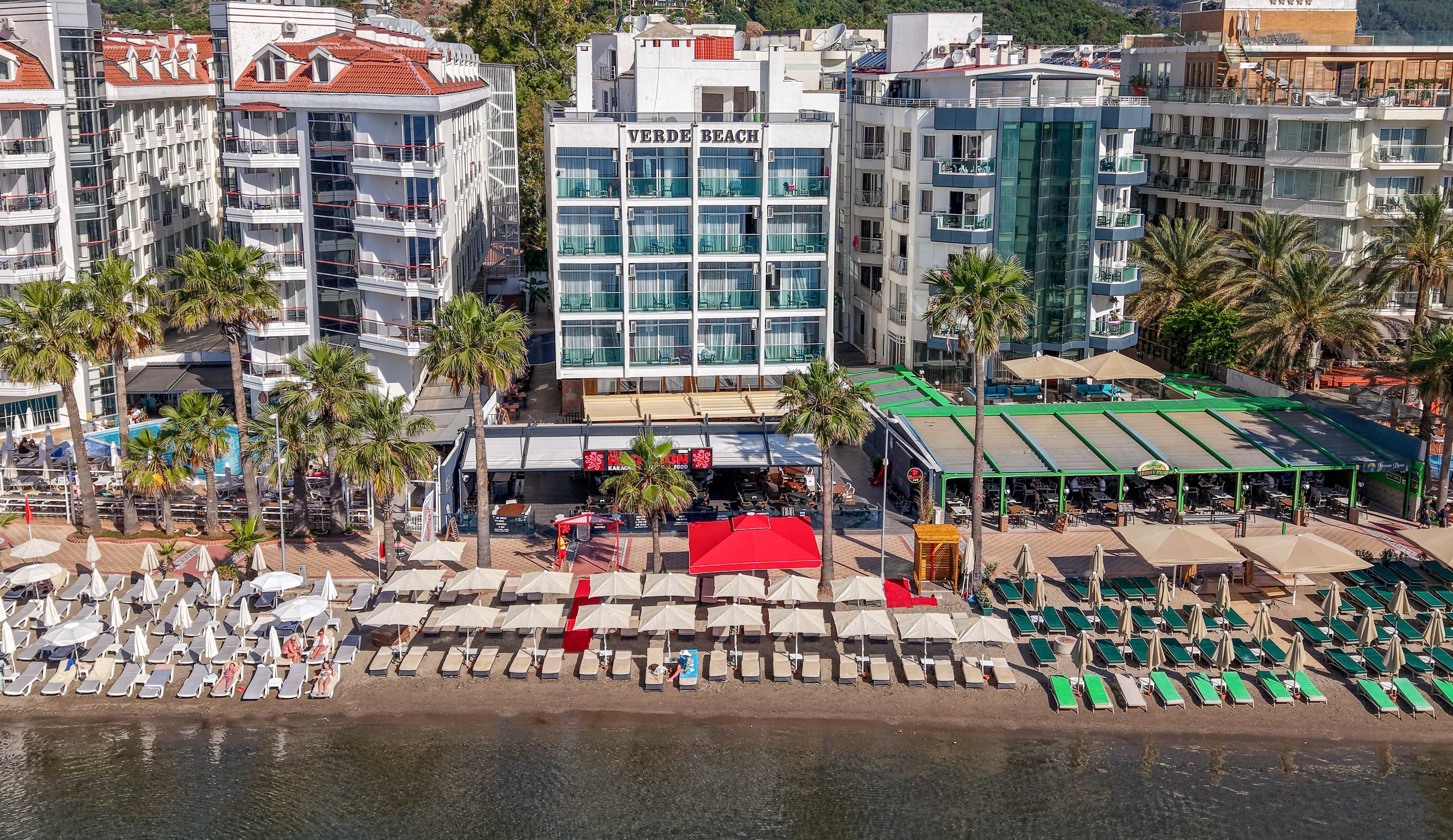 A beachfront hotel labeled VERDE BEACH faces a sandy shoreline lined with sun loungers and umbrellas. The aerial view shows several multi-story buildings, palm trees, and a covered terrace area beside the water. Neatly arranged rows of white and green loungers extend toward the calm sea, with a few people relaxing near the shore. The scene suggests a popular coastal resort destination under clear daylight.