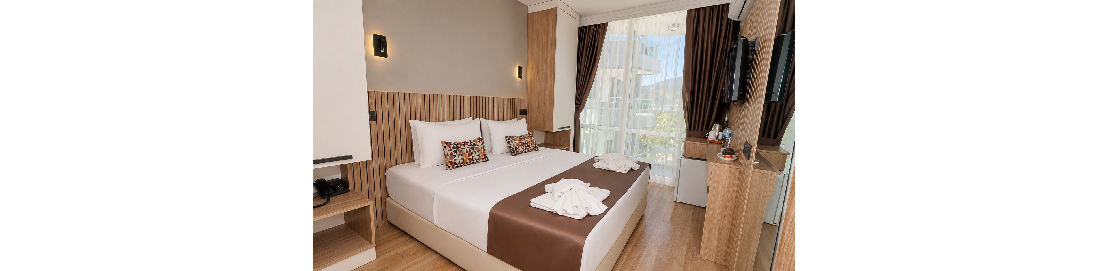 A contemporary hotel room features a neatly made double bed with white linens and a brown runner. Two patterned accent pillows and towel art on the bed add decorative touches. Built-in wooden furniture, including bedside tables, a desk, and shelving, create a clean and organized look. Floor-to-ceiling windows with sheer curtains and dark drapes open to a balcony with an outdoor view, while a wall-mounted TV and minibar complete the cozy space.