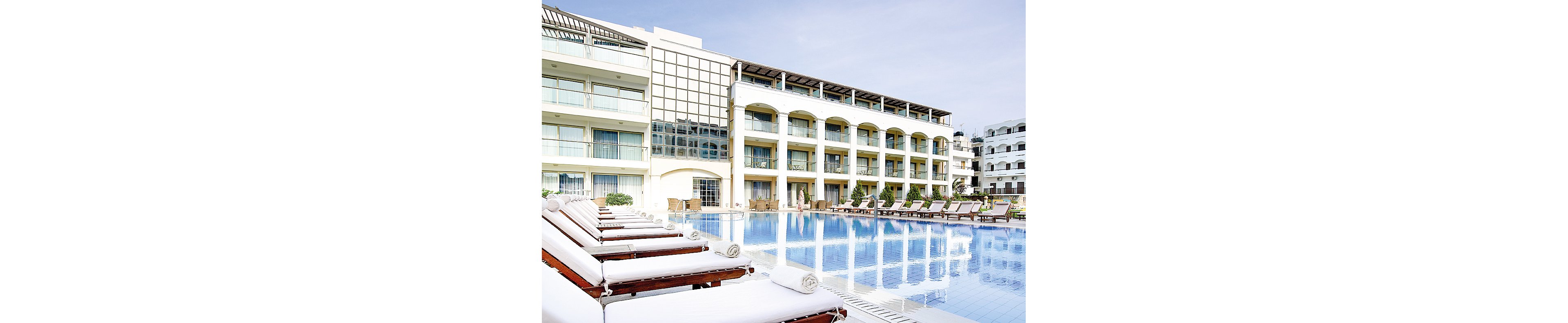 A serene view of a luxury resort featuring a large outdoor pool surrounded by white lounge chairs. The setting includes a modern multi-story building with balconies and glass windows. The clear blue sky enhances the tranquil atmosphere, perfect for relaxation and leisure.
