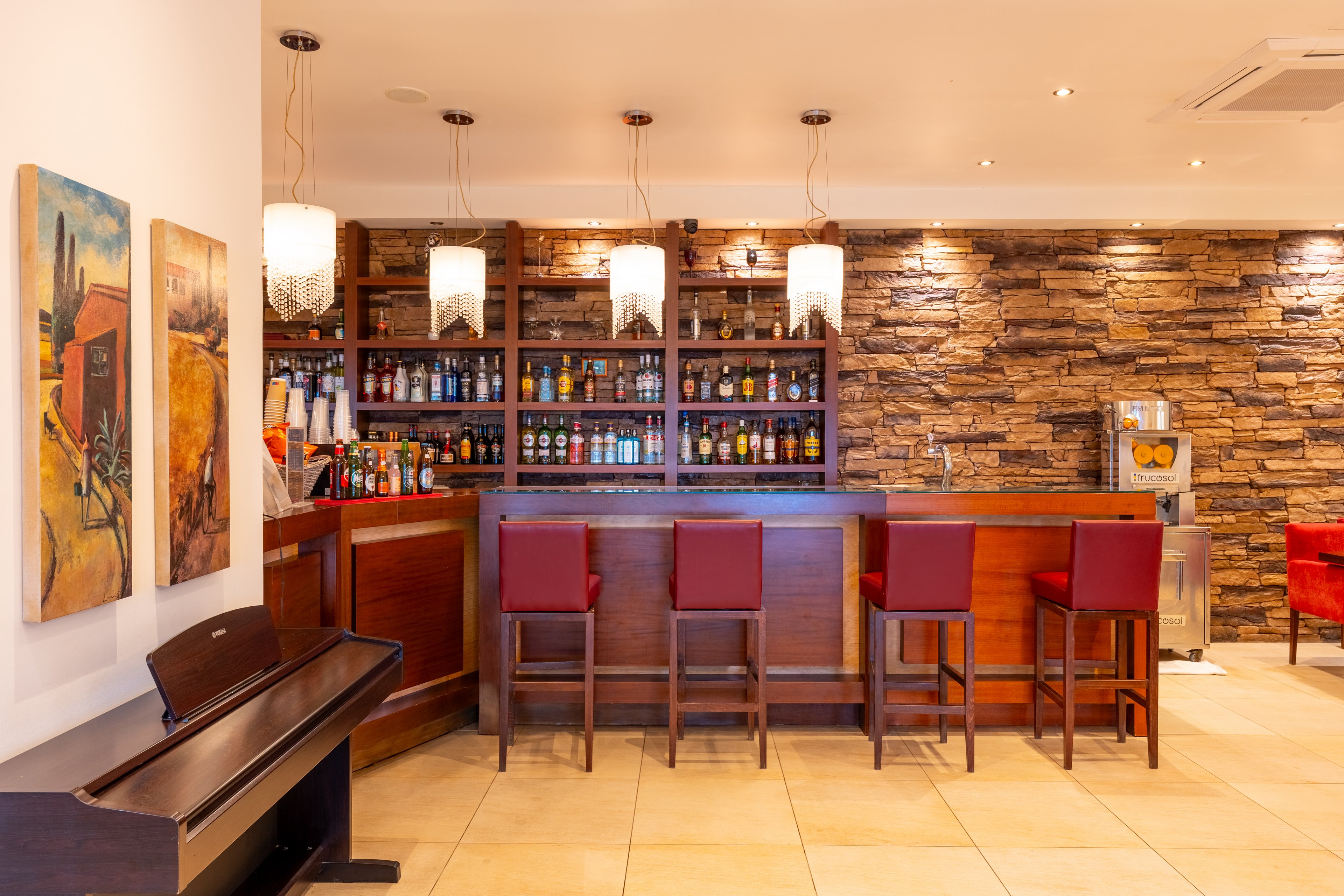Warmly lit bar interior features a wooden counter with four red bar stools and a stone-clad back wall. Shelves behind the bar display an assortment of bottles and glassware, creating a well-stocked, inviting atmosphere. Pendant lights hang above the counter, while a piano and colorful paintings add artistic touches to the space. The tiled floor and clean, modern ceiling complete the contemporary lounge setting.