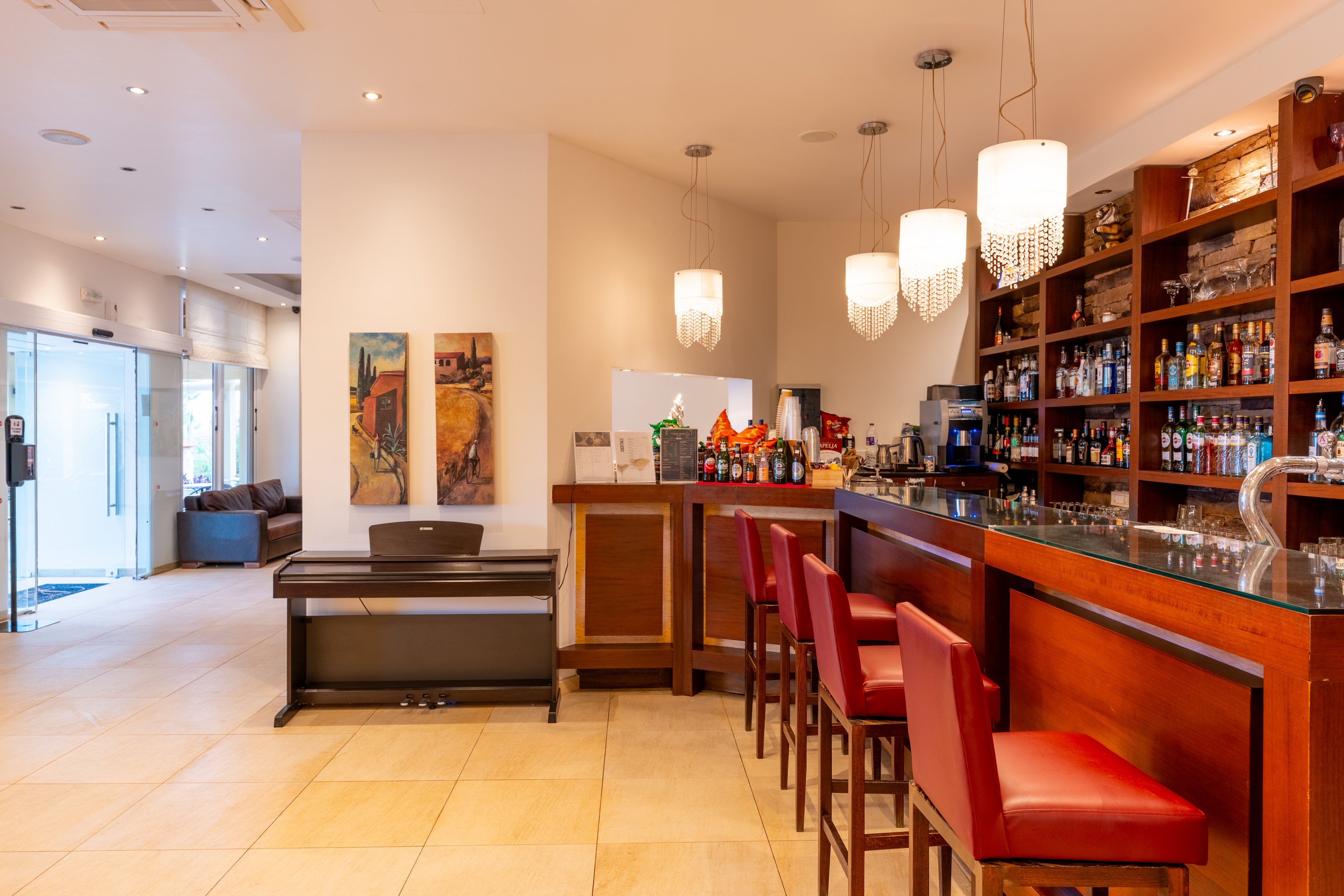 Bright contemporary bar area featuring a polished counter and red upholstered bar stools. Shelves behind the bar display a wide variety of bottles and glassware. Modern pendant lights hang from the ceiling, illuminating the warm wood finishes and tiled floor. Artwork on the walls and a piano in the background add to the stylish, welcoming atmosphere.