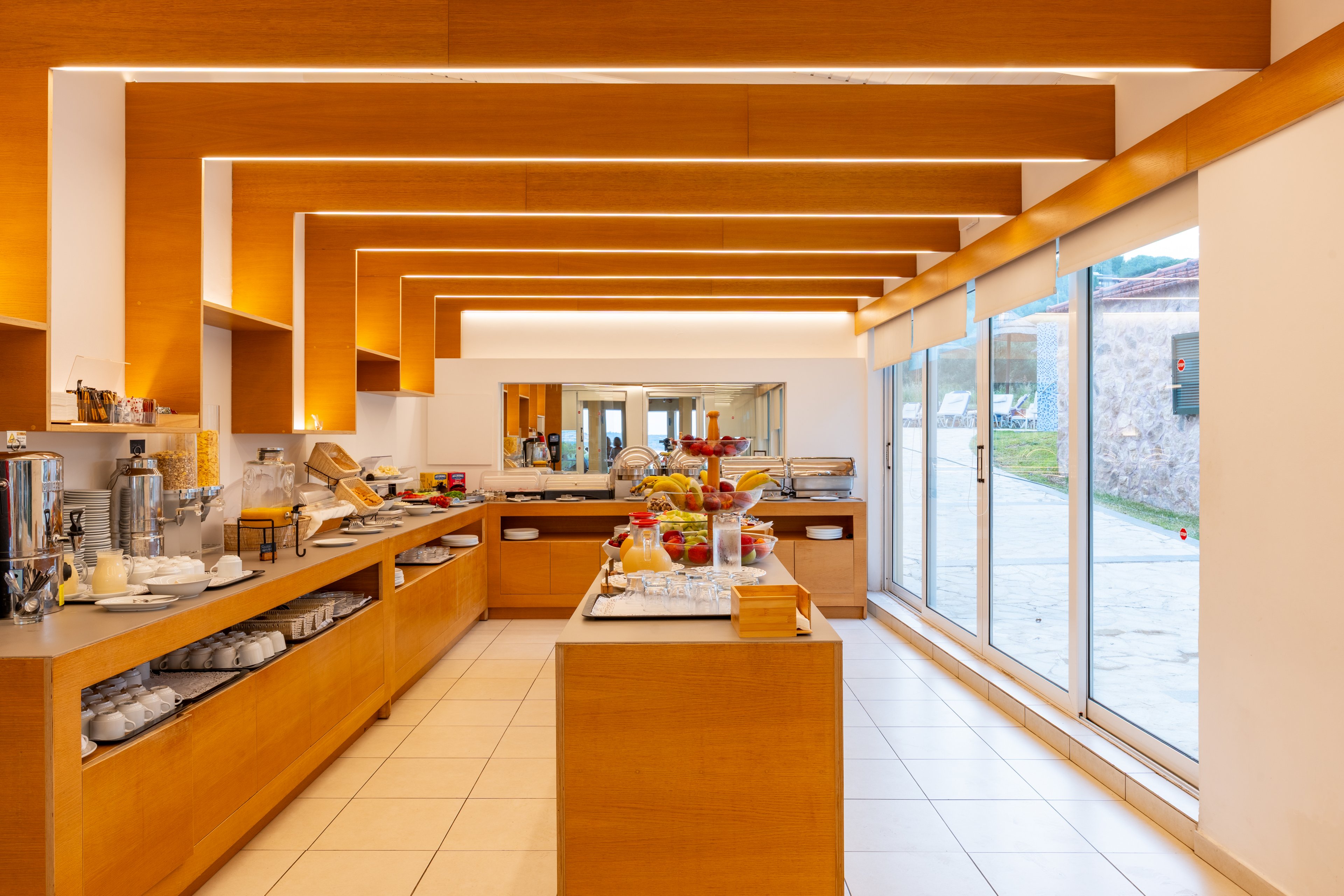 A contemporary self-service breakfast buffet is arranged on sleek wooden counters in a bright hotel dining area. Various foods, beverages, and tableware are neatly displayed, with coffee urns and juice dispensers visible on the left. Large floor-to-ceiling windows on the right let in natural daylight and reveal an outdoor stone wall and greenery. Warm wood tones, clean lines, and recessed ceiling lighting create an inviting, modern atmosphere.
