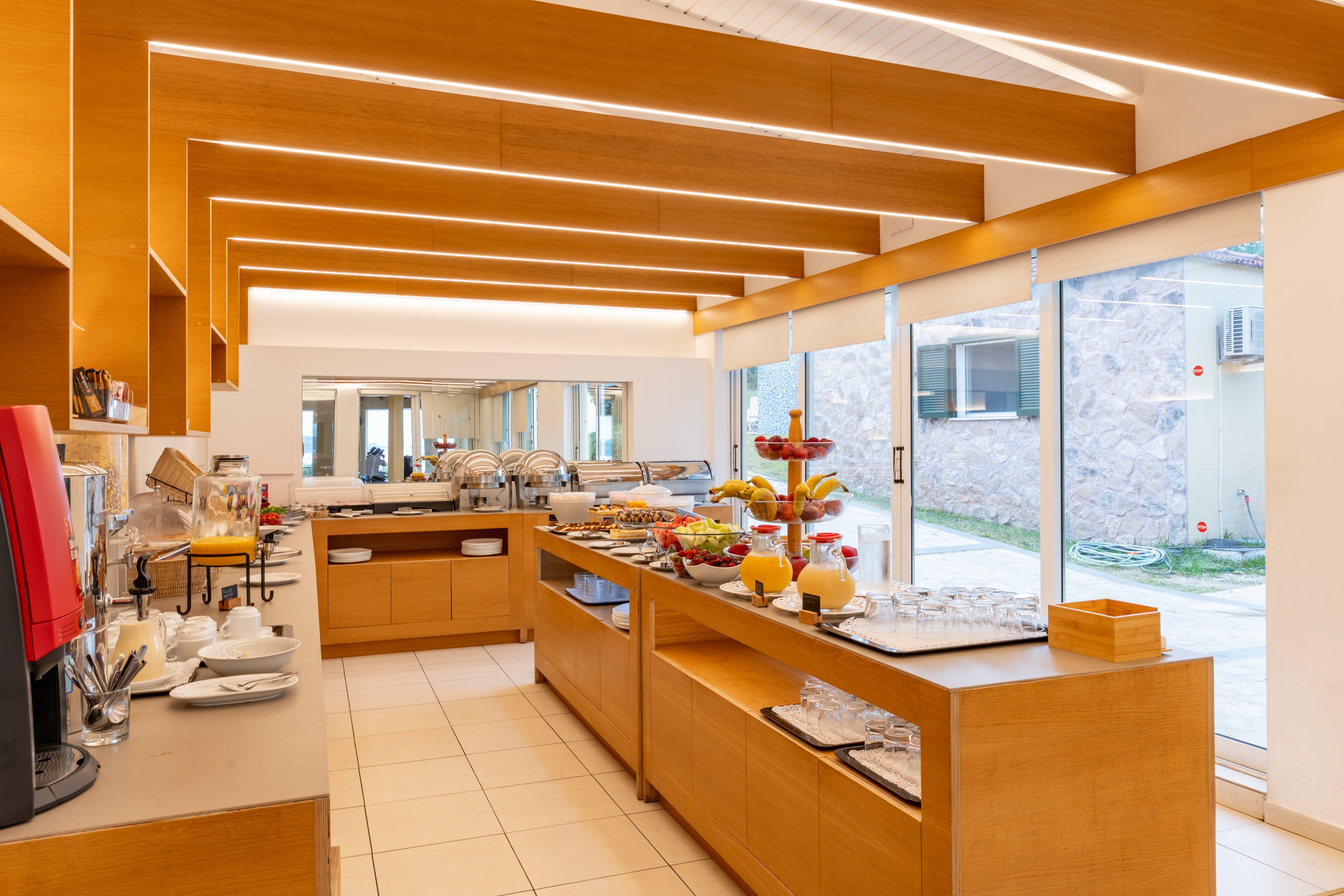 A bright self-service breakfast buffet is arranged on wooden counters in a modern dining area. Various foods, including fruits, pastries, cereals, and hot dishes, are neatly displayed in trays and stands. Large floor-to-ceiling windows let in natural light and reveal an exterior stone wall and garden. Clean lines, warm wood tones, and recessed ceiling lighting create a contemporary, inviting atmosphere.