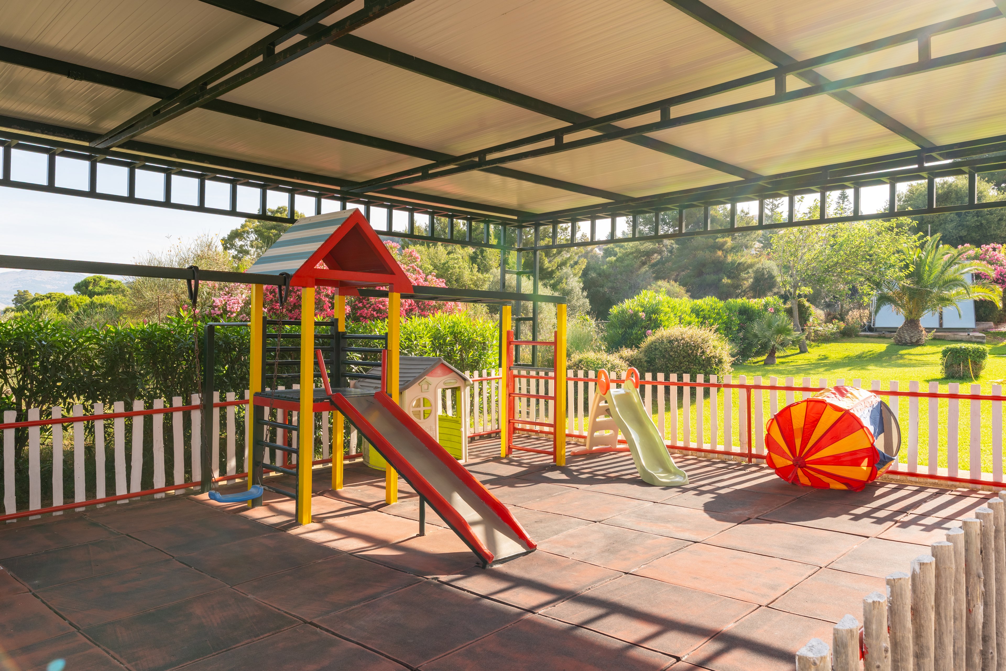 A small covered playground features bright red and yellow play structures on a tiled floor. A slide, climbing frame, and playhouse sit within a white picket fence enclosure. Sunlight streams in from the open sides, revealing a lush green garden and trees in the background. The scene suggests a safe, cheerful play area for children in an outdoor setting.