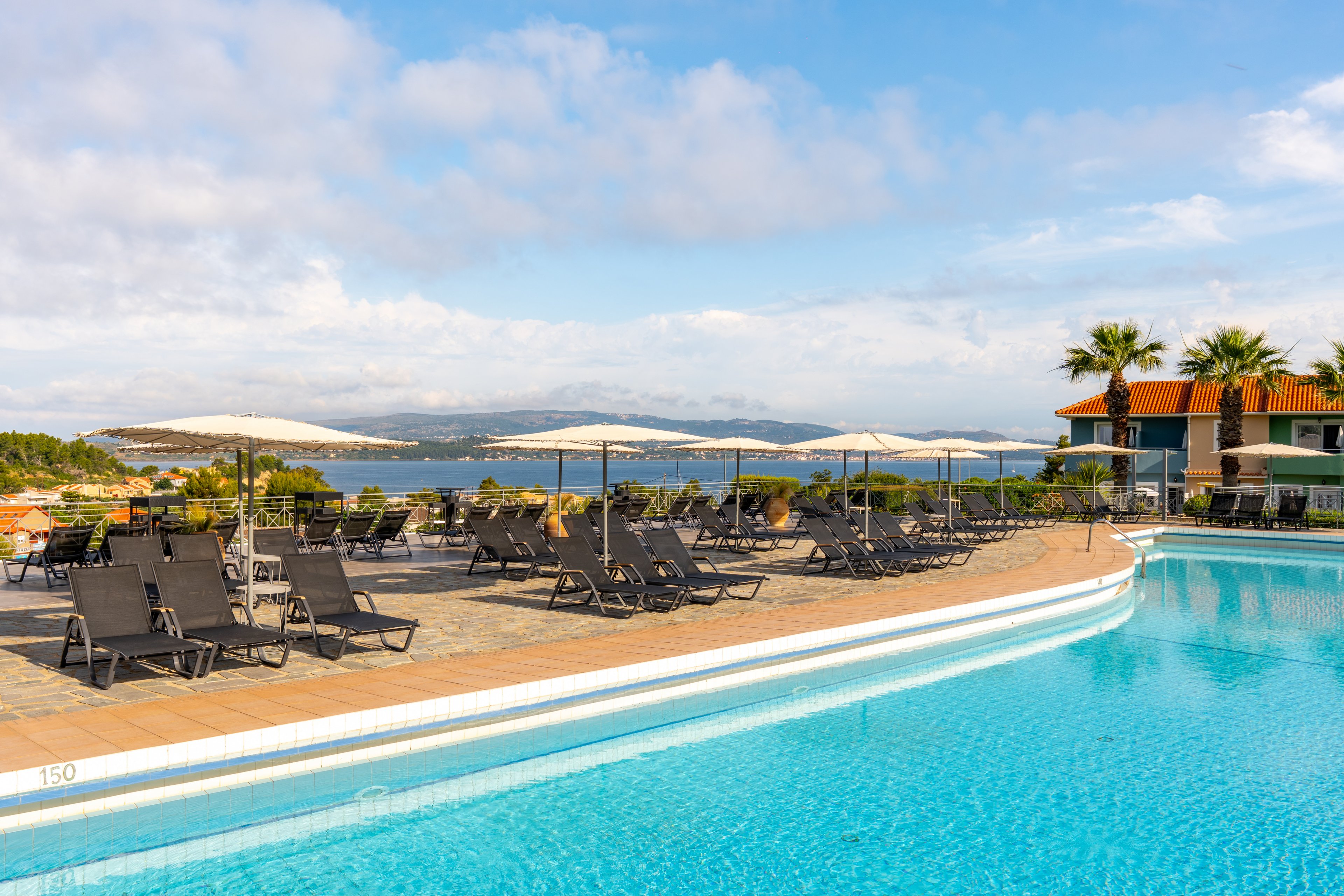 An outdoor swimming pool at a seaside resort is lined with black sun loungers and large white parasols. The clear blue water of the pool reflects the bright sky, while palm trees and tiled buildings frame the scene. In the background, a calm bay and distant hills create a relaxing coastal atmosphere. The area appears quiet and empty, suggesting an inviting, tranquil vacation setting.