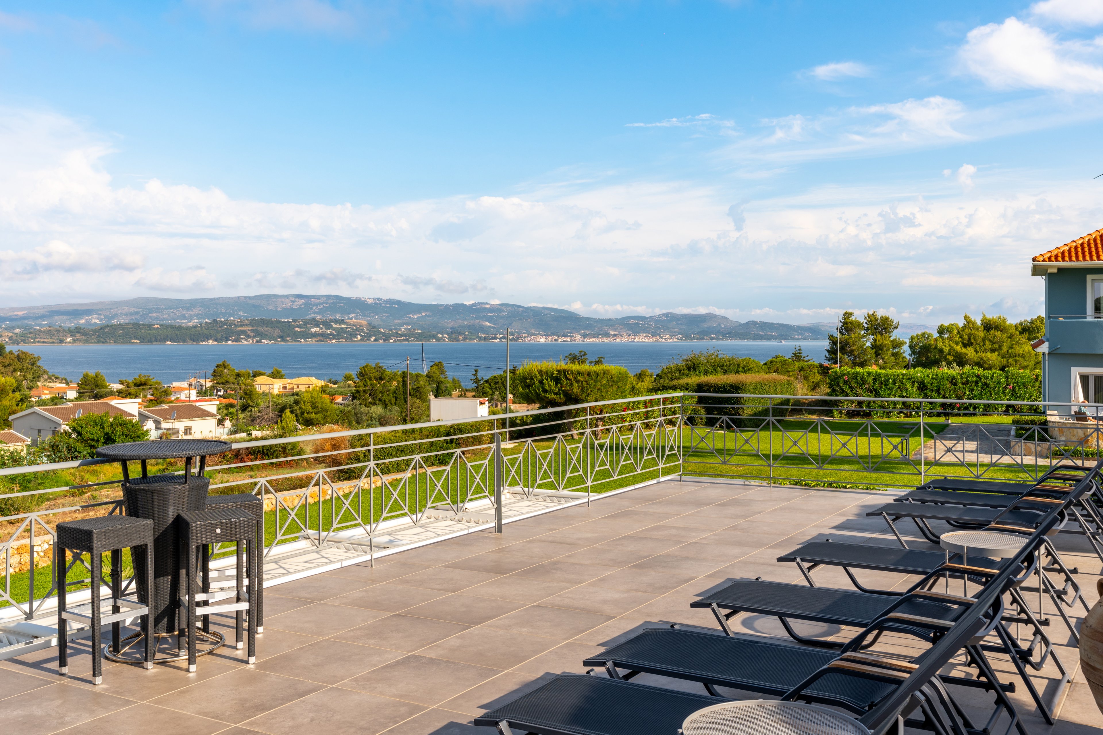 A spacious terrace features multiple dark lounge chairs arranged in rows facing a scenic coastal view. A tall table with bar stools stands near the railing, inviting guests to relax and enjoy the landscape. Beyond the terrace, houses, greenery, and distant hills surround a calm body of water under a bright blue sky with scattered clouds. The overall scene suggests a peaceful vacation or resort setting in warm weather.