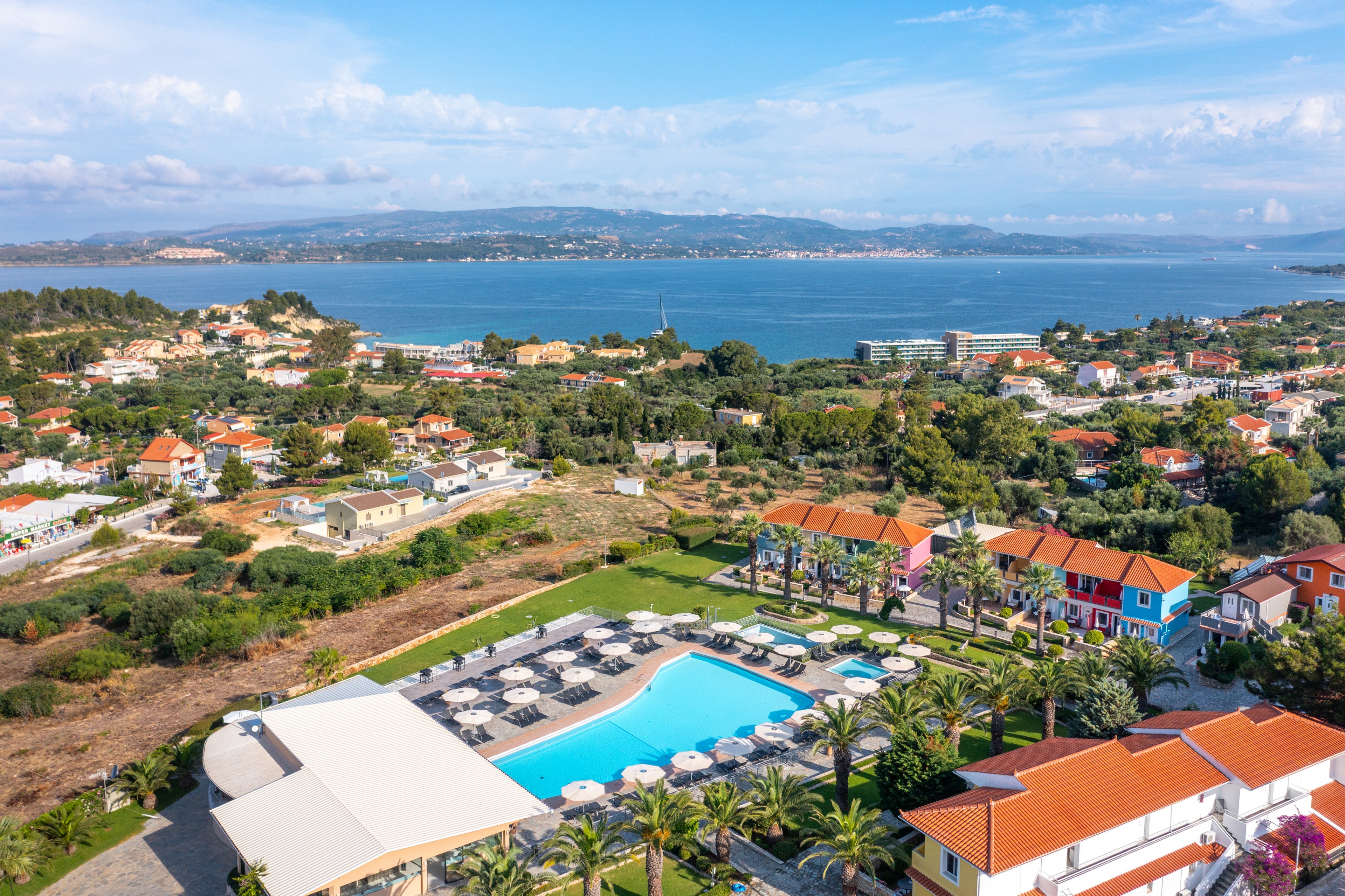 Aerial view of a colorful coastal resort featuring a large outdoor swimming pool surrounded by sun loungers and palm trees. The property sits among red-roofed buildings and greenery, with a calm bay and distant hills in the background. Clear blue water in the pool and sea contrasts with the bright rooftops and lush landscape, suggesting a sunny vacation destination. The scene conveys relaxation, leisure, and scenic seaside living.