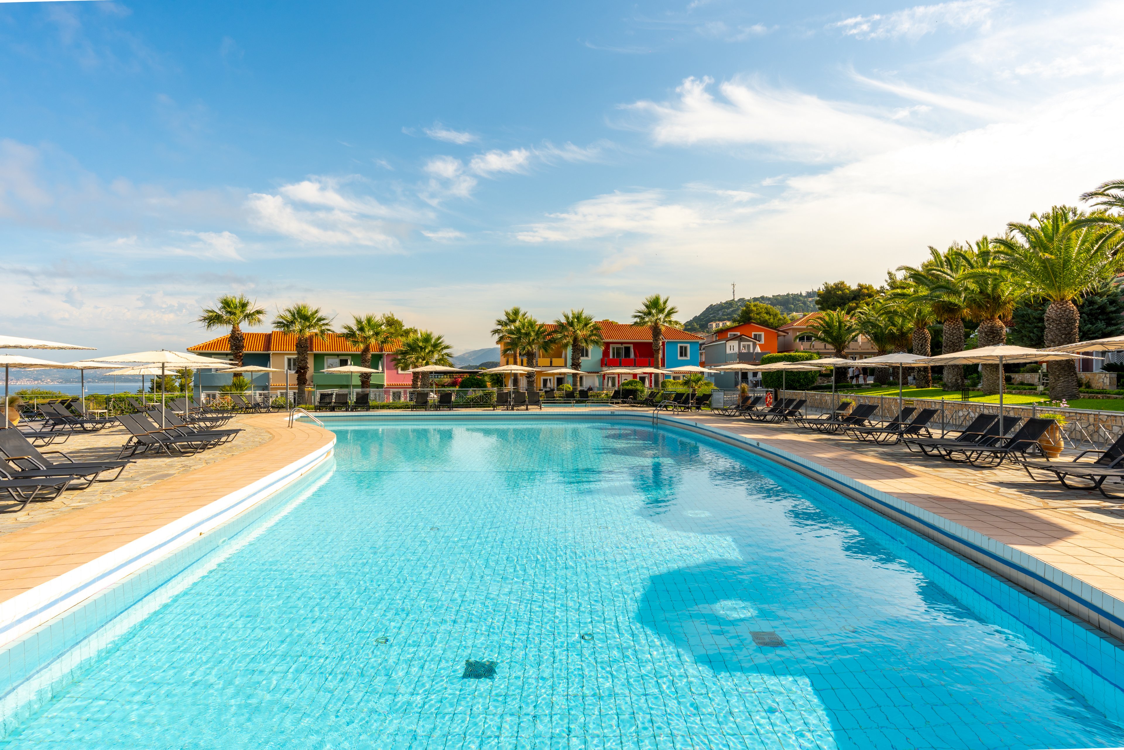 A large outdoor swimming pool stretches toward a row of colorful villas in a seaside resort. Sun loungers and umbrellas line the pool deck, suggesting a relaxing vacation setting. Palm trees frame the scene under a bright blue sky with scattered clouds. The calm water and vibrant buildings create a cheerful, inviting holiday atmosphere.