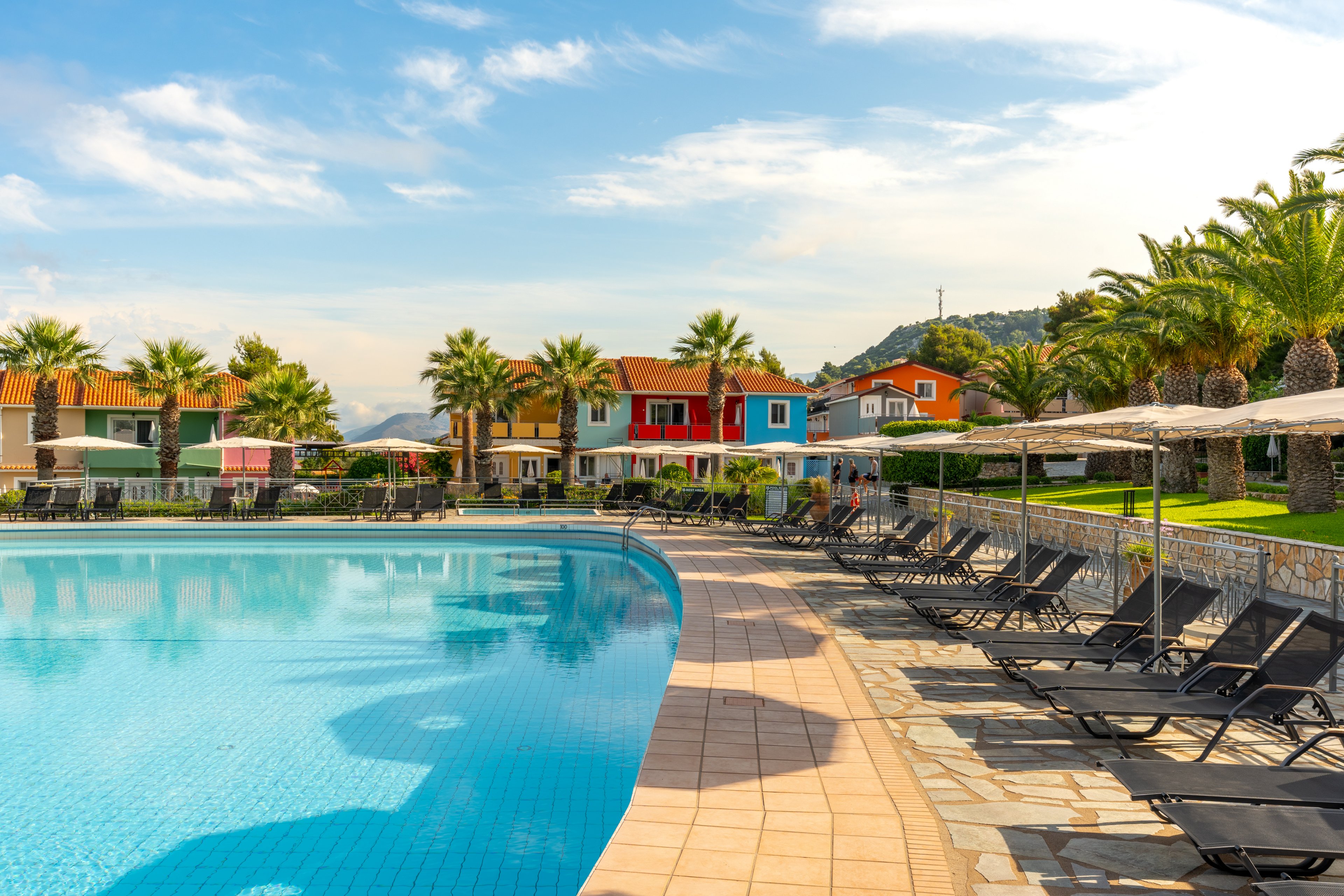 A large outdoor swimming pool sits in front of a row of colorful resort villas. Numerous black lounge chairs and umbrellas line the tiled poolside terrace, suggesting a relaxing vacation setting. Palm trees and manicured lawns frame the scene, with hills and blue sky in the background. The bright daylight and clear water create a warm, inviting atmosphere ideal for leisure and travel.