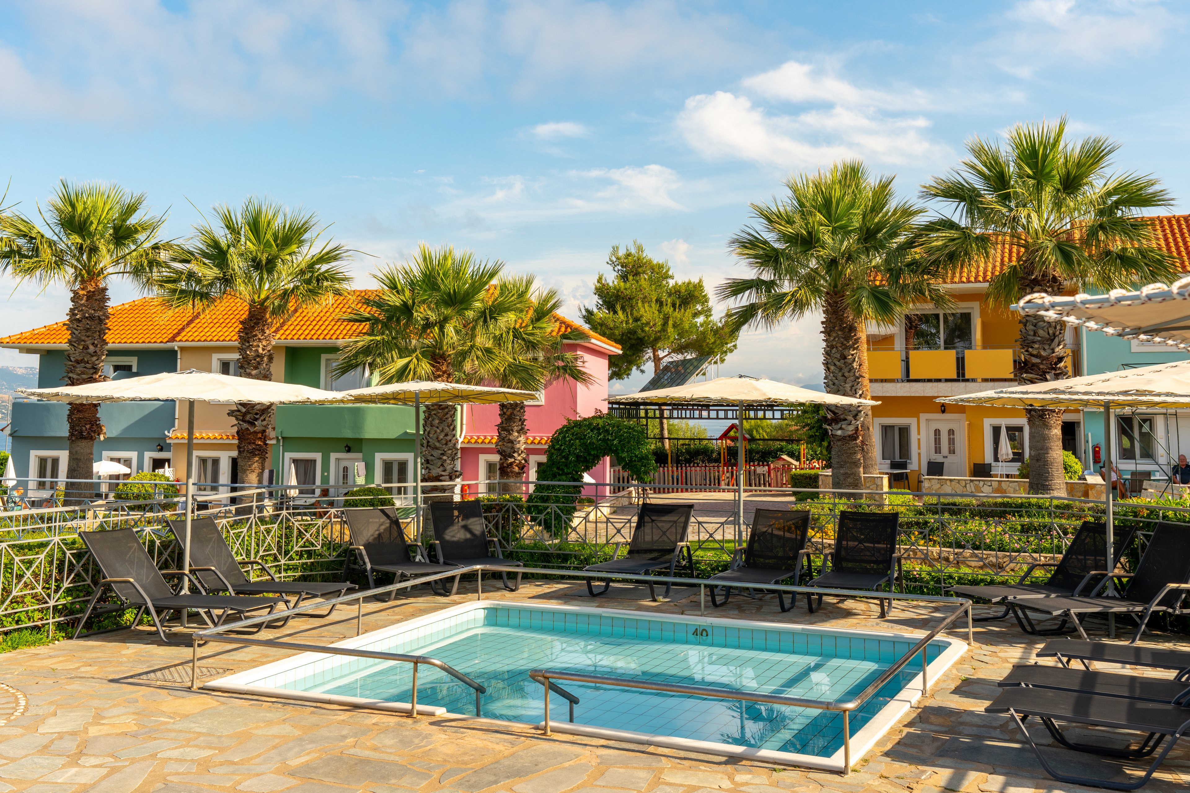 A small rectangular swimming pool is surrounded by black lounge chairs and white sun umbrellas in a sunny courtyard. Tall palm trees line the pool area, adding a tropical feel to the scene. In the background, brightly painted houses in yellow, pink, blue, and orange create a vibrant, cheerful atmosphere. The clear blue sky and lush greenery suggest a warm vacation destination.