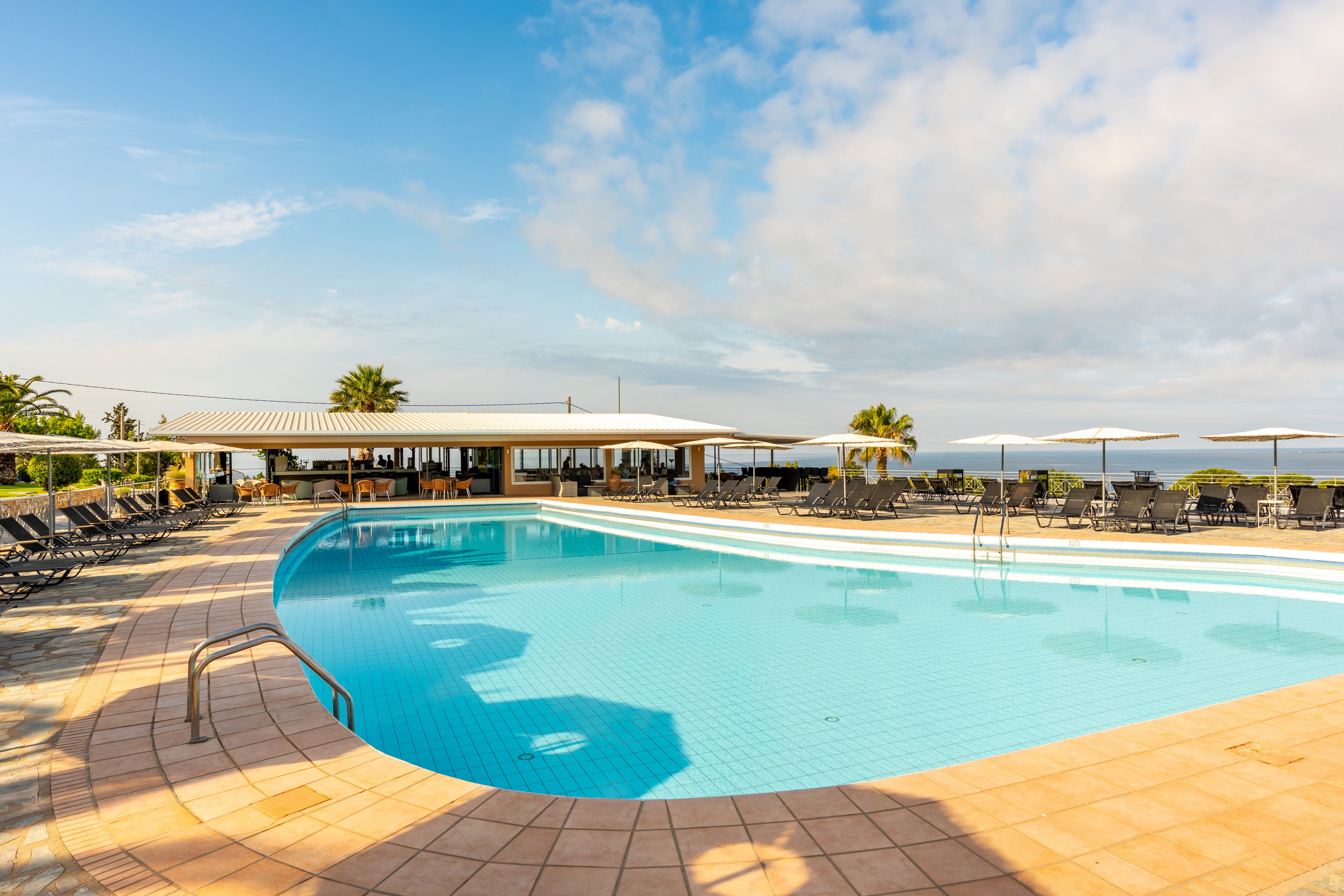 Outdoor resort swimming pool with clear blue water is shown under a bright sunny sky. Lounge chairs and umbrellas surround the poolside terrace, creating a relaxing vacation atmosphere. A low modern building with open seating stands behind the pool, and palm trees add a tropical touch. The distant horizon reveals a calm sea, enhancing the tranquil coastal setting.