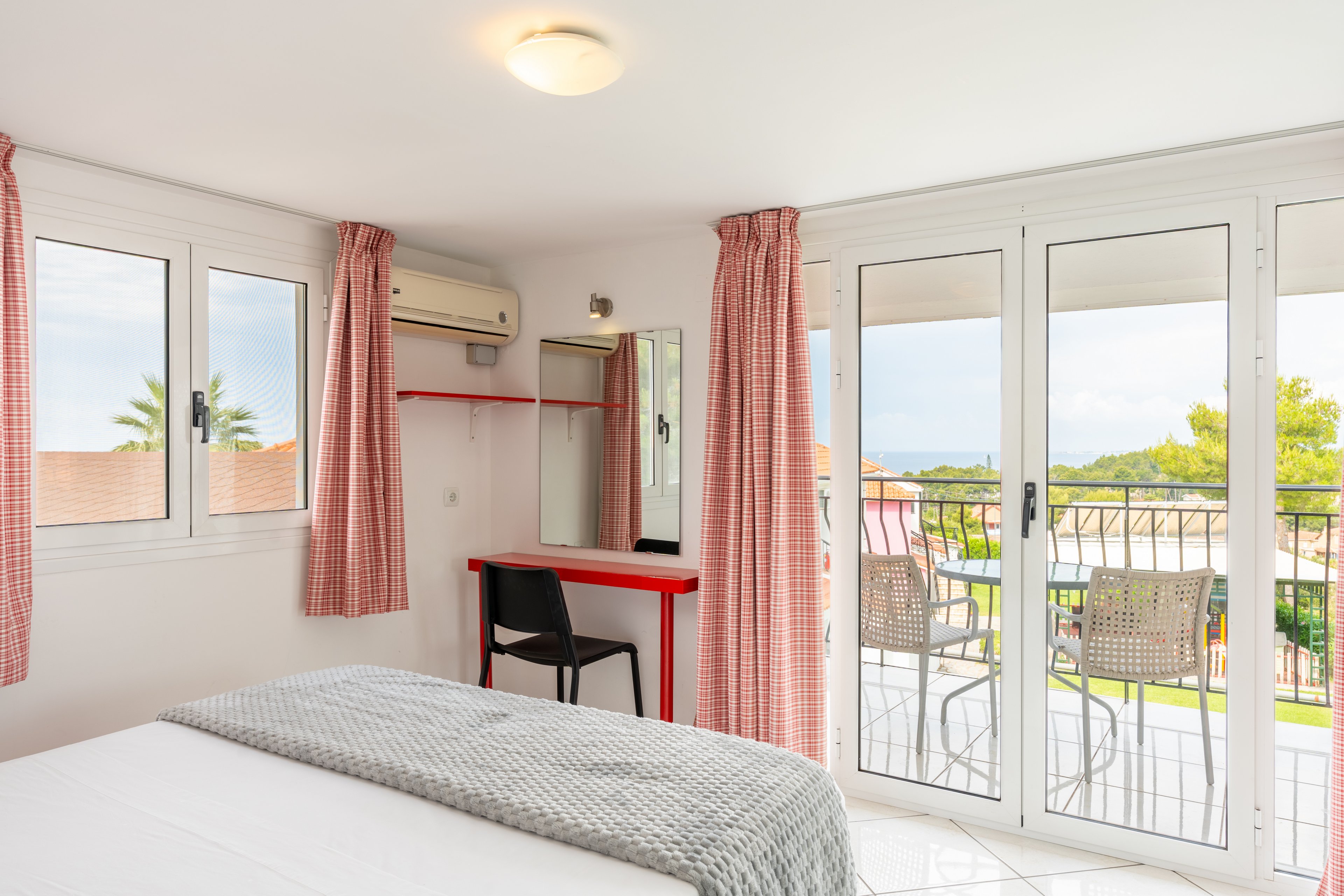 A sunlit hotel bedroom features a neatly made bed with a light gray throw blanket. Large glass doors open to a balcony with outdoor chairs and a scenic view of greenery and rooftops. Pink patterned curtains frame both the balcony doors and side windows, adding a soft touch of color. A small desk with a mirror and black chair sits against the wall beneath an air conditioning unit, creating a simple workspace.