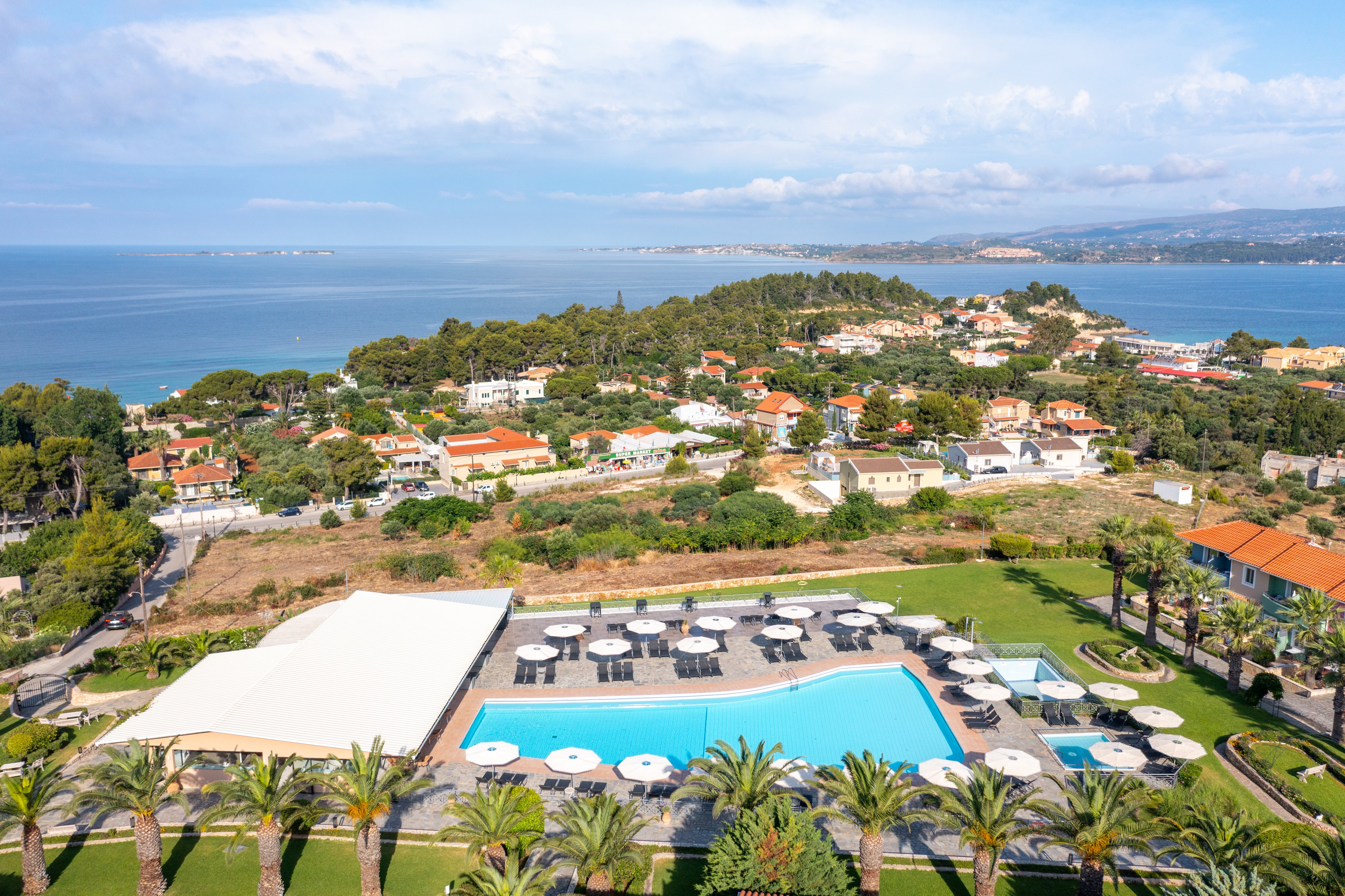 A large resort swimming pool is surrounded by sun loungers and umbrellas in a landscaped garden with palm trees. The scene overlooks a coastal village with houses and greenery stretching toward the shoreline. Beyond the buildings, the calm blue sea extends to the horizon under a partly cloudy sky. The elevated viewpoint highlights the contrast between the manicured resort grounds and the natural coastal landscape.
