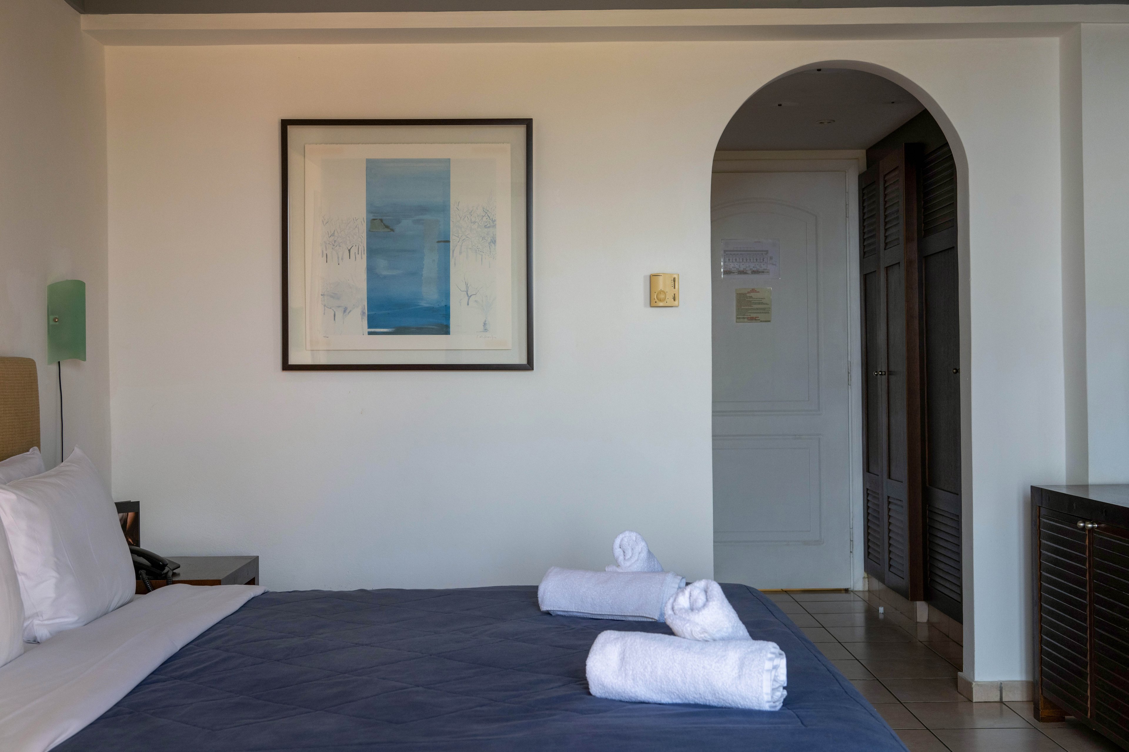 A neatly made bed with a dark blue cover and white pillows is the main focus of this hotel room. Three rolled white towels are placed on the bed, suggesting a prepared and welcoming space. The room features a framed artwork on the wall, a white door through an arched doorway, and built-in closets. Soft natural light and neutral colors create a calm, minimalist atmosphere suitable for relaxation or travel accommodation.