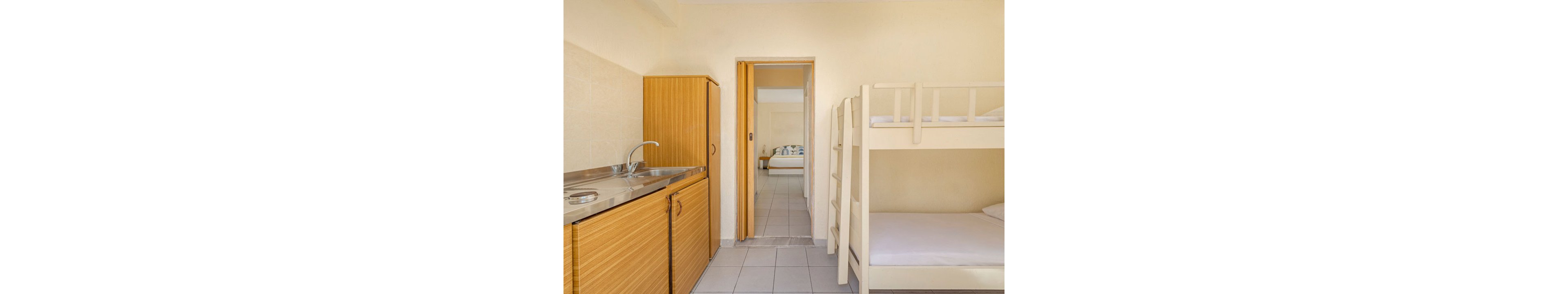 Small studio room features a simple kitchenette with a stainless steel sink and wooden cabinets. A white bunk bed with clean bedding stands against the wall, emphasizing efficient use of space. The tiled floor and neutral walls create a bright, minimal aesthetic. Through the open doorway, another room with a double bed is visible, suggesting a connected apartment layout.