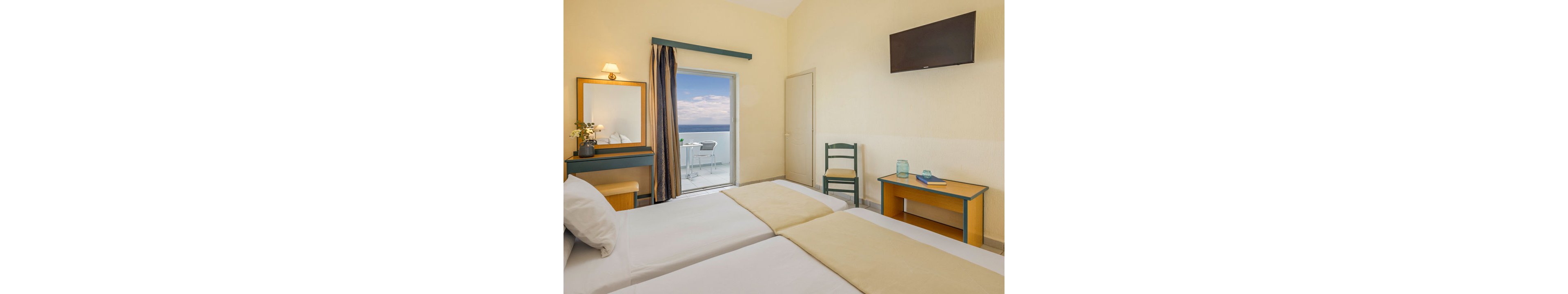 A tidy hotel room features two single beds with light beige runners and white linens. The space includes a wooden dressing table with a mirror, a small desk with decorative items, and a simple chair. A wall-mounted flat-screen television hangs above the desk, and a doorway opens to a balcony with white furniture overlooking the sea and sky. Soft yellow walls and teal accents create a calm, coastal atmosphere.