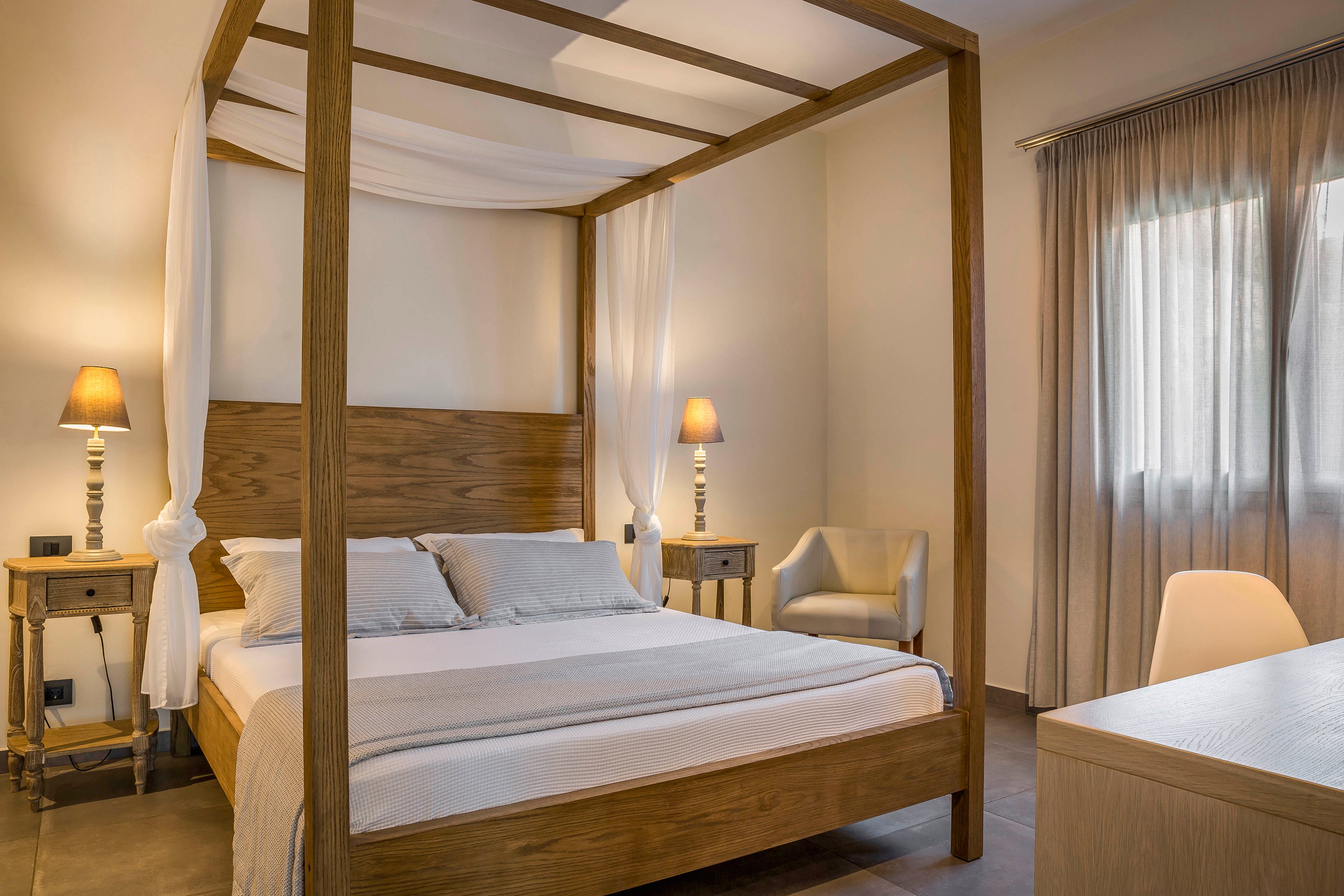 A contemporary bedroom features a wooden four-poster canopy bed neatly made with neutral bedding and pillows. Warm bedside lamps on matching wooden nightstands create a soft, inviting glow. A light-colored armchair sits in the corner near a window dressed with sheer curtains, allowing gentle daylight into the room. The overall decor is minimalist, with clean lines and a calm, relaxing atmosphere.