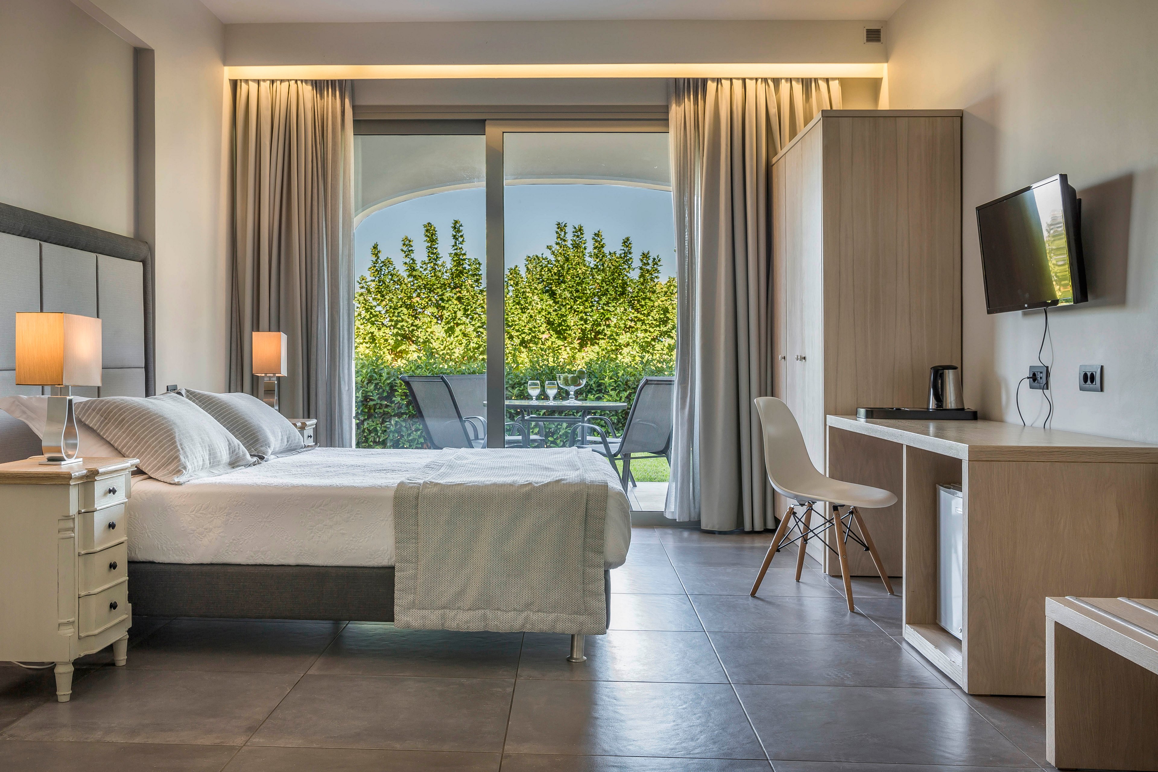 A contemporary hotel bedroom features a neatly made double bed with soft white linens and bedside lamps. Large sliding glass doors open to a balcony with outdoor chairs and a lush green garden view. The room includes a wall-mounted television, desk area with chair, and a small refrigerator beneath the counter. Neutral tones, clean lines, and soft lighting create a calm and inviting atmosphere.