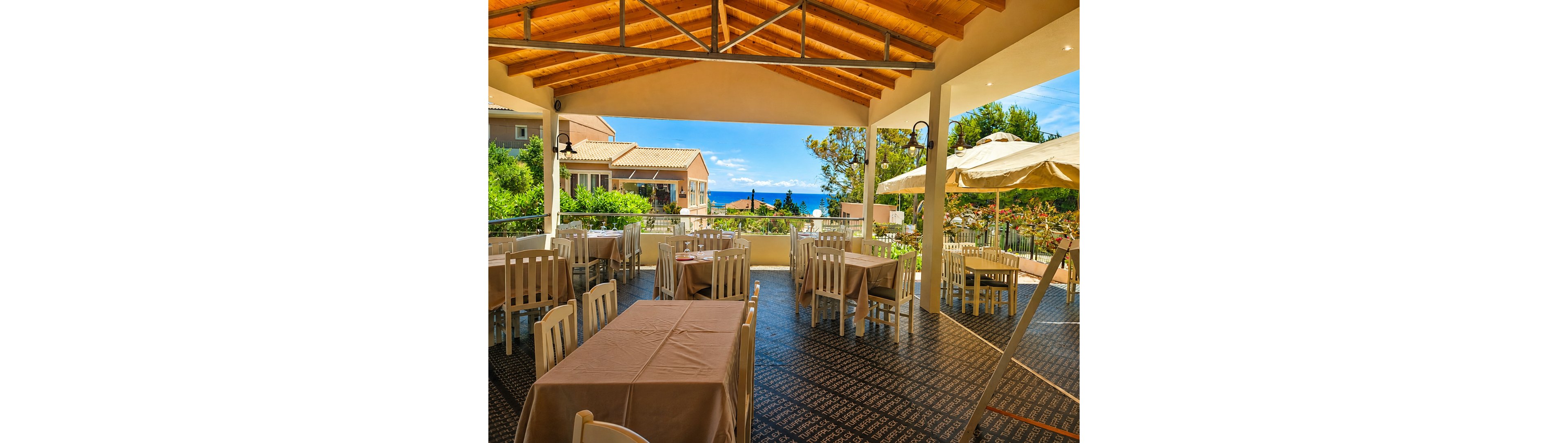 An open-air restaurant terrace features neatly arranged tables covered with beige tablecloths and white wooden chairs. The setting overlooks lush greenery, nearby buildings, and a distant view of the blue sea under a bright sky. A large patio umbrella provides shade on one side, while a wooden ceiling with exposed beams shelters the main dining area. Warm sunlight and vibrant colors create a relaxed, inviting Mediterranean atmosphere.