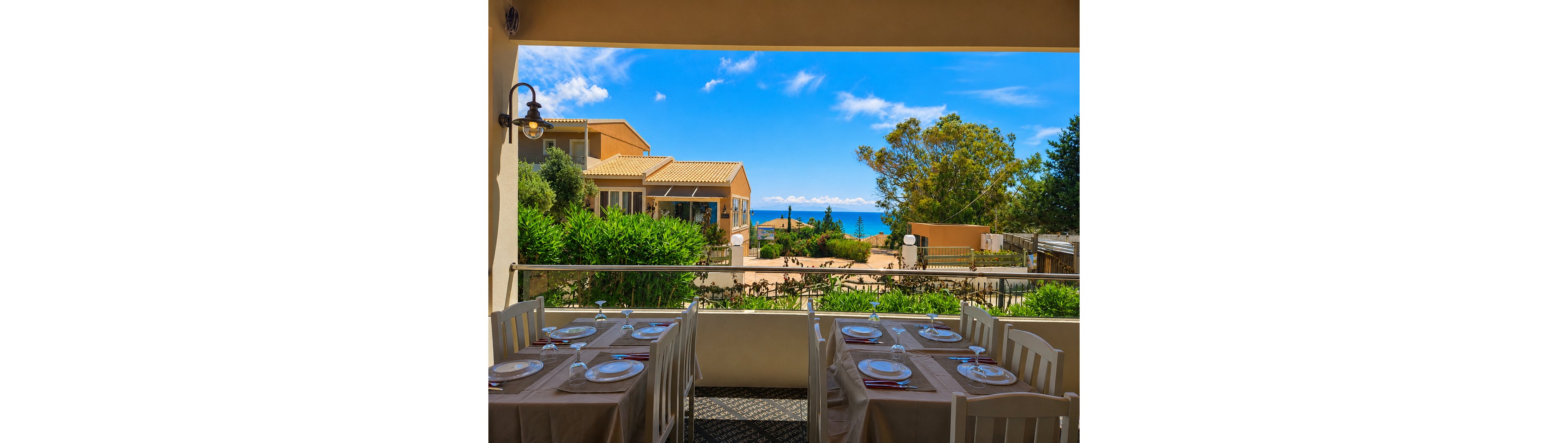 An outdoor restaurant terrace is set with neatly arranged tables and chairs, each covered with beige tablecloths and white plates. The terrace overlooks a courtyard with lush greenery and light-colored buildings. Beyond the buildings, the blue sea stretches out under a bright sky with scattered clouds. The scene suggests a relaxed coastal dining experience in warm, sunny weather.