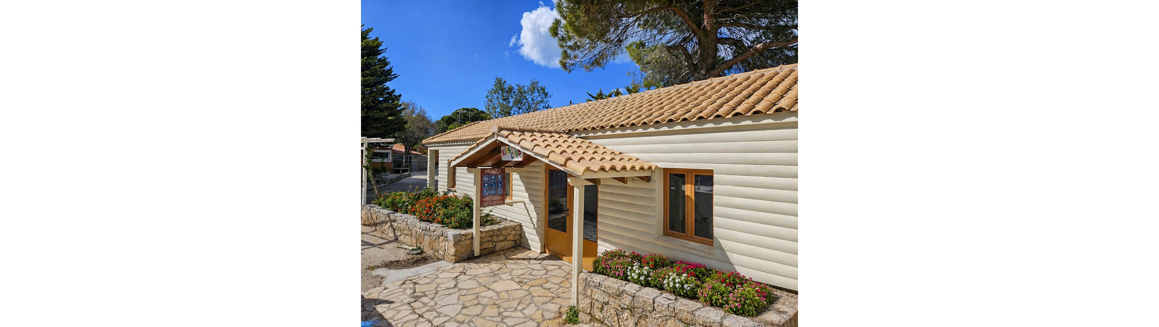 A single-story wooden building with a tiled roof stands in a sunny outdoor setting. The entrance is framed by a small covered porch and large glass doors. Colorful flowers line the stone pathway and low stone wall in front of the structure. Tall trees and a bright blue sky with scattered clouds create a pleasant, inviting atmosphere.