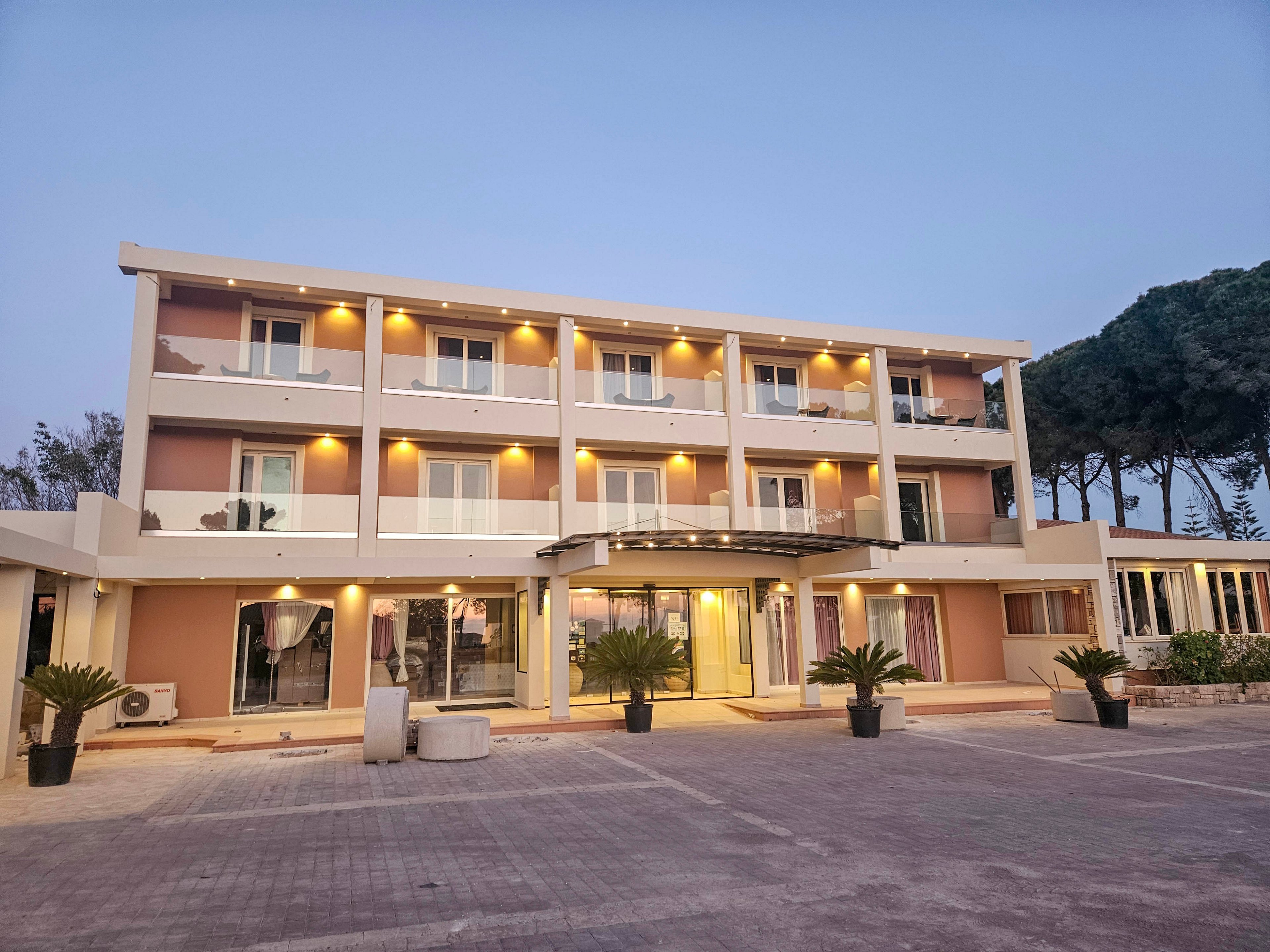 A modern three-story hotel building is photographed at dusk with warm exterior lights illuminating each balcony. The structure features clean lines, large windows, and a central glass-covered entrance. Potted plants and small trees decorate the front area, which opens onto a wide paved courtyard. Tall trees and a clear evening sky form the backdrop, creating a calm and inviting atmosphere.