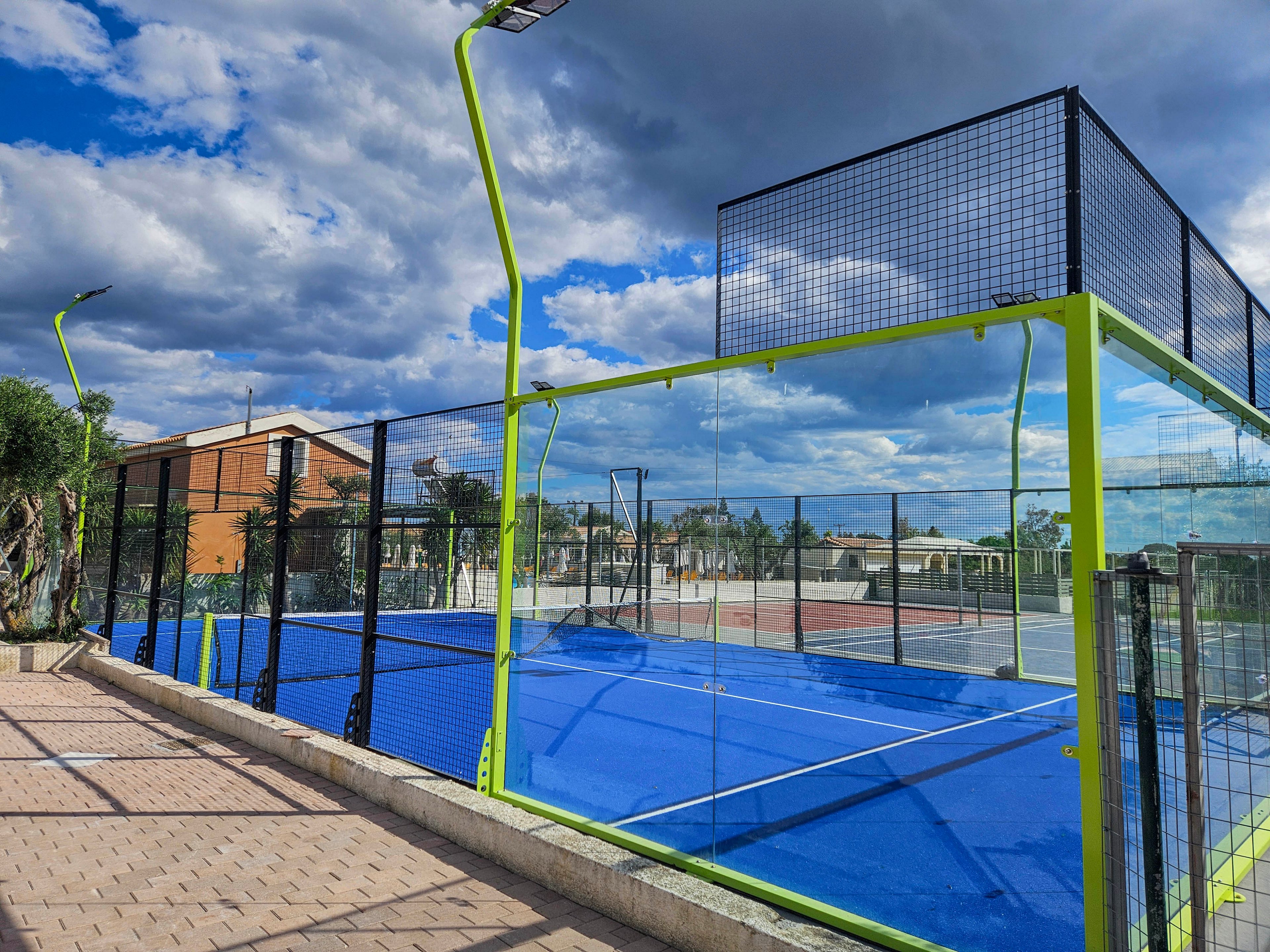 A bright blue padel court is enclosed by tall glass and metal fencing with neon green posts. The court is located outdoors beside a paved walkway and landscaped greenery. Residential buildings and additional sports courts are visible in the background under a dramatic cloudy sky. The scene is empty, emphasizing the clean lines of the court and the vibrant contrast between the blue surface and the sky.