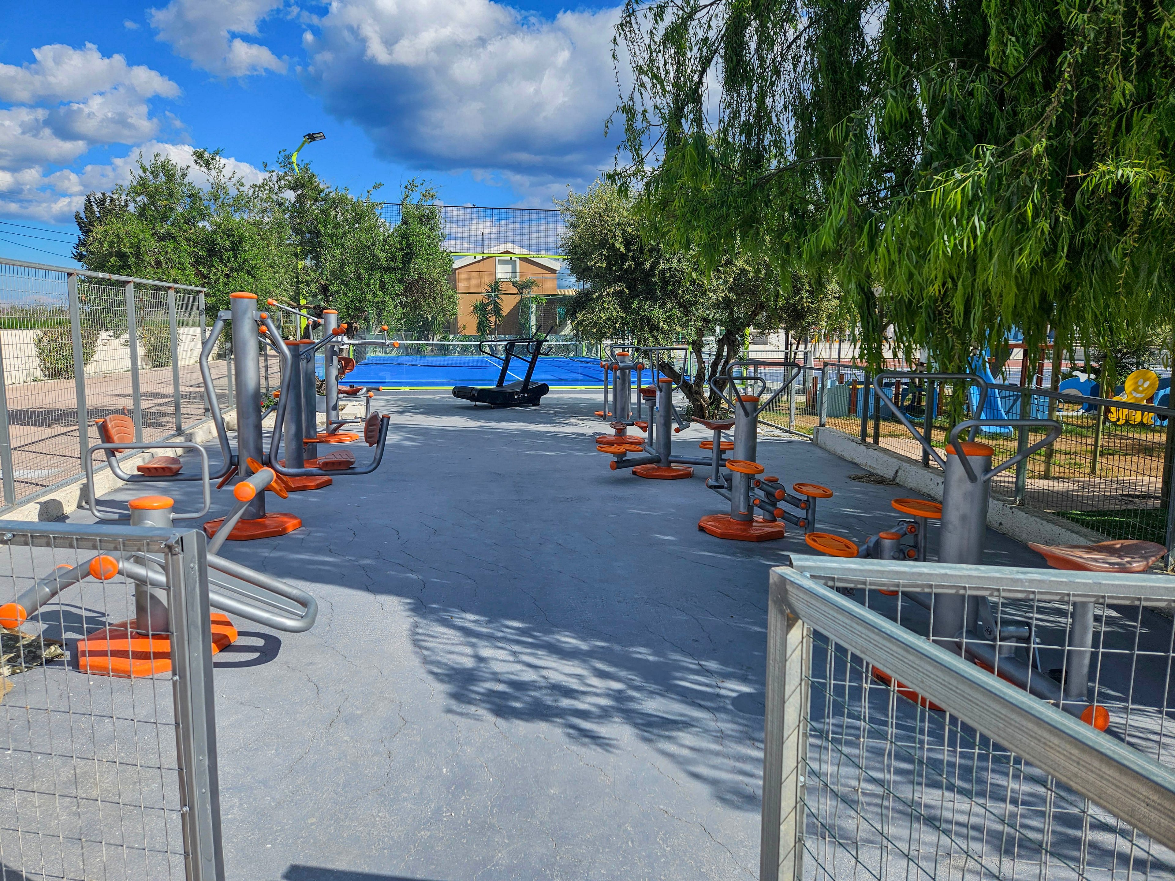 An open-air fitness area features multiple metal exercise machines with orange seats and accents. The equipment is arranged on a smooth concrete surface, enclosed by metal fencing. Trees provide shade around the perimeter, and a blue structure or pool is visible in the background. The scene is brightly lit under a partly cloudy sky, suggesting a pleasant day for outdoor exercise.