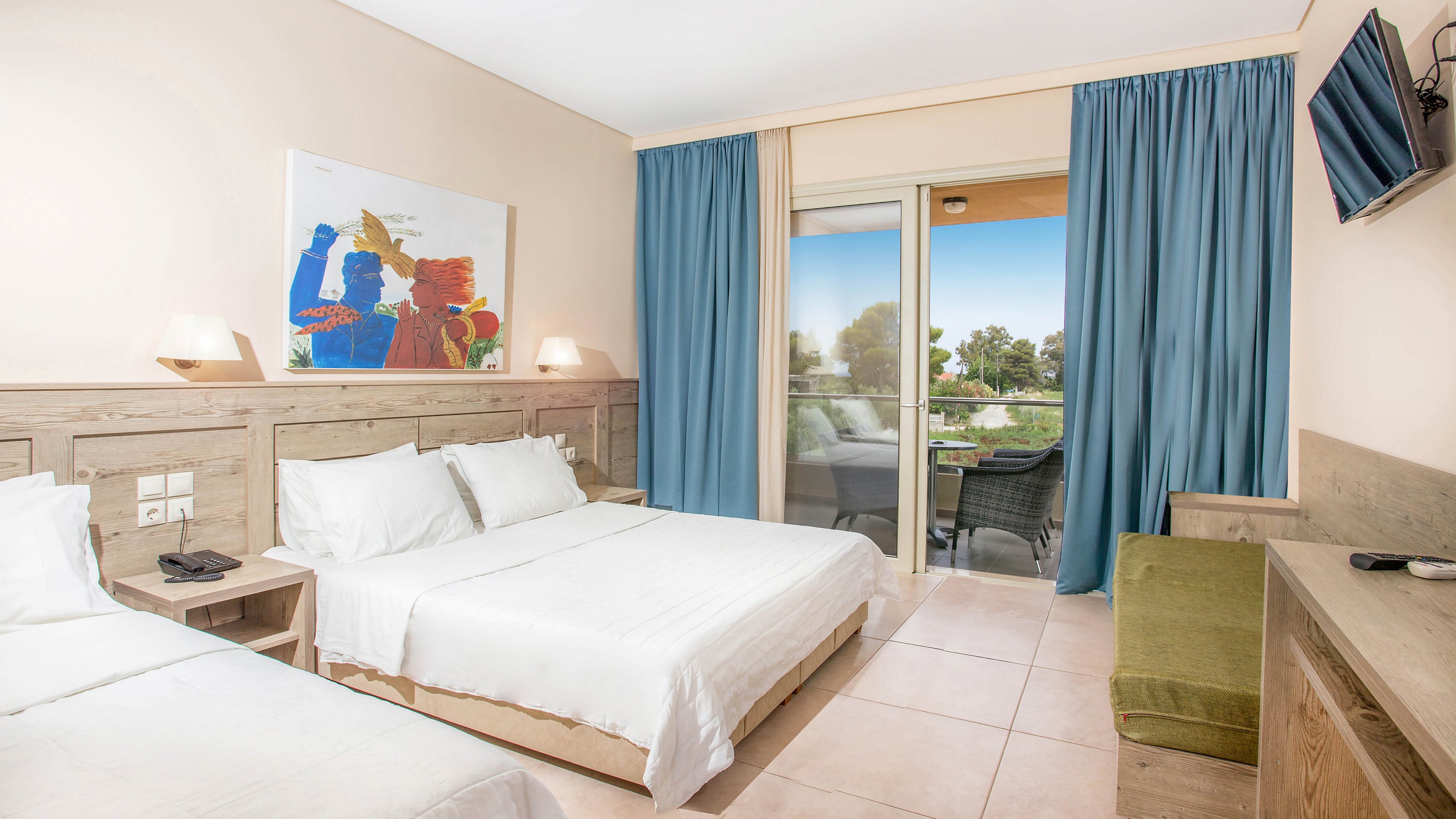 A modern hotel room features a double bed and a single bed with crisp white linens against light wood furnishings. Blue curtains frame a glass door that opens to a balcony overlooking greenery and an outdoor area. A colorful artwork hangs above the headboard, while a wall-mounted television and desk complete the interior. The space is clean, bright, and inviting, with natural light enhancing the neutral tones.
