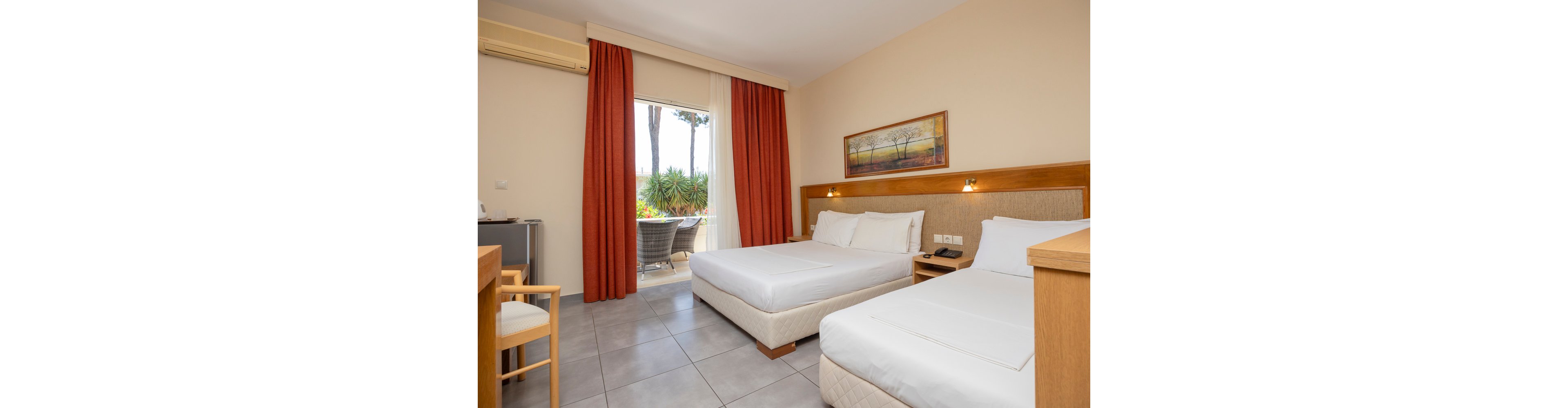A clean, modern hotel room features two neatly made beds with white linens and a wooden headboard. The space includes a small desk, chair, mini fridge, and wall-mounted air conditioner. Warm wall lights and a landscape painting add a cozy touch to the neutral decor. Large glass doors with red curtains open to a small balcony with outdoor seating and greenery visible outside.