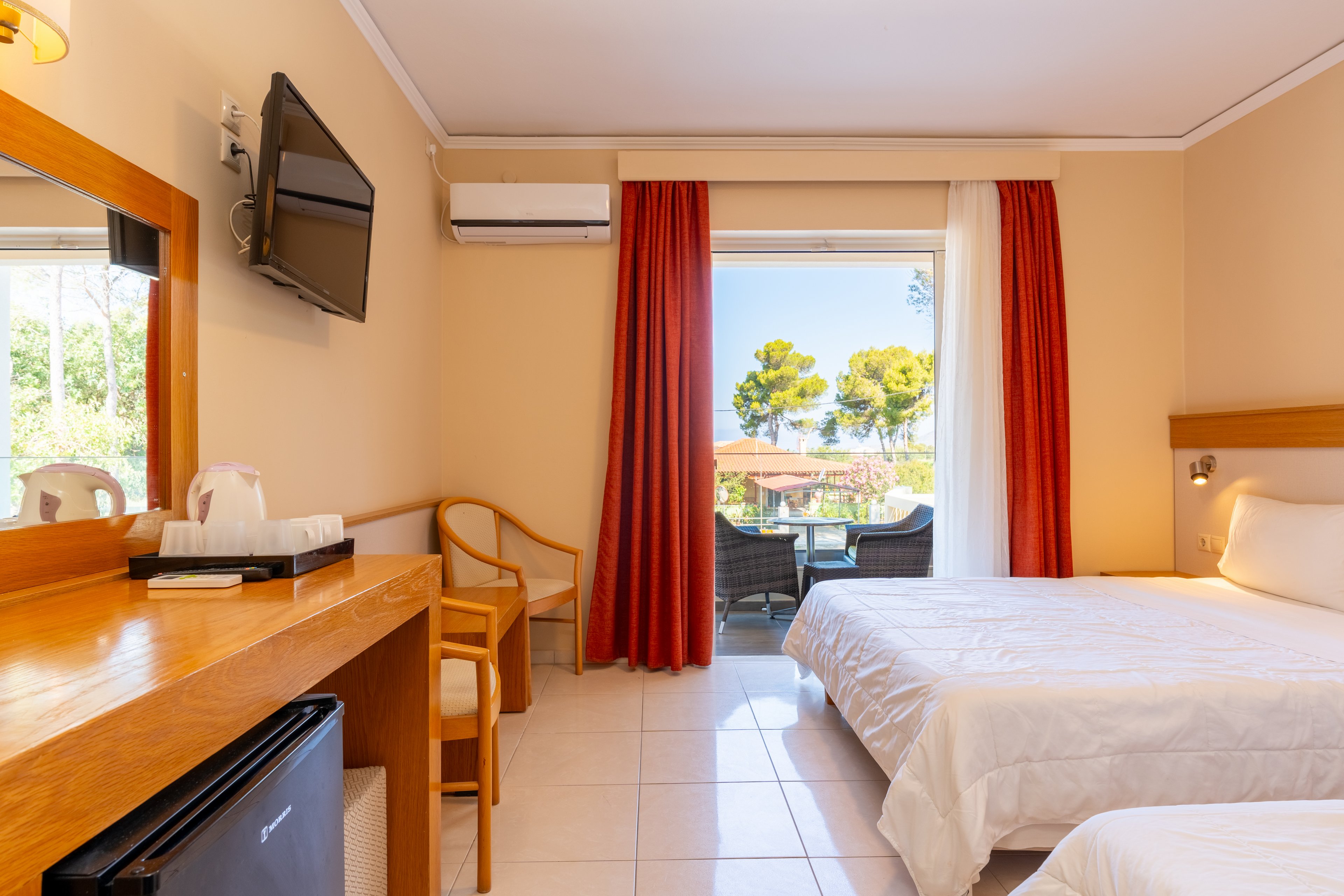 A tidy hotel room features two white-covered beds and a wooden desk with a chair and mini fridge. A wall-mounted television and air conditioning unit are visible above the desk area. Large red curtains frame a glass door leading to a balcony with outdoor chairs and a scenic view of trees and buildings. Natural light fills the space, creating a warm and inviting atmosphere.