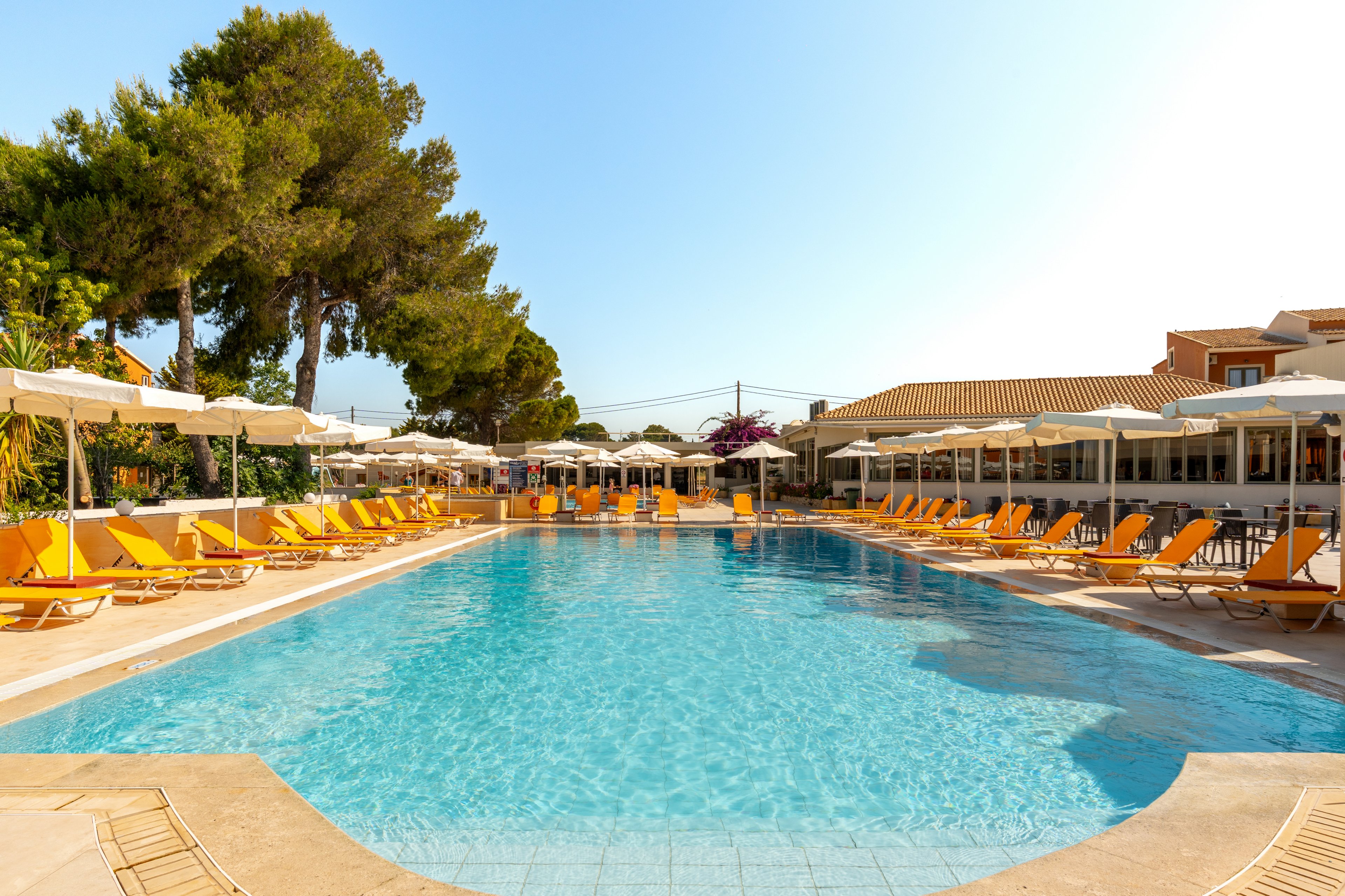 A large outdoor swimming pool is surrounded by rows of yellow sun loungers and white umbrellas at a resort. The scene is set on a bright, clear day with blue sky overhead. Trees and low buildings frame the pool area, creating a relaxed holiday atmosphere. The calm water and orderly seating suggest a peaceful, well-maintained vacation destination.