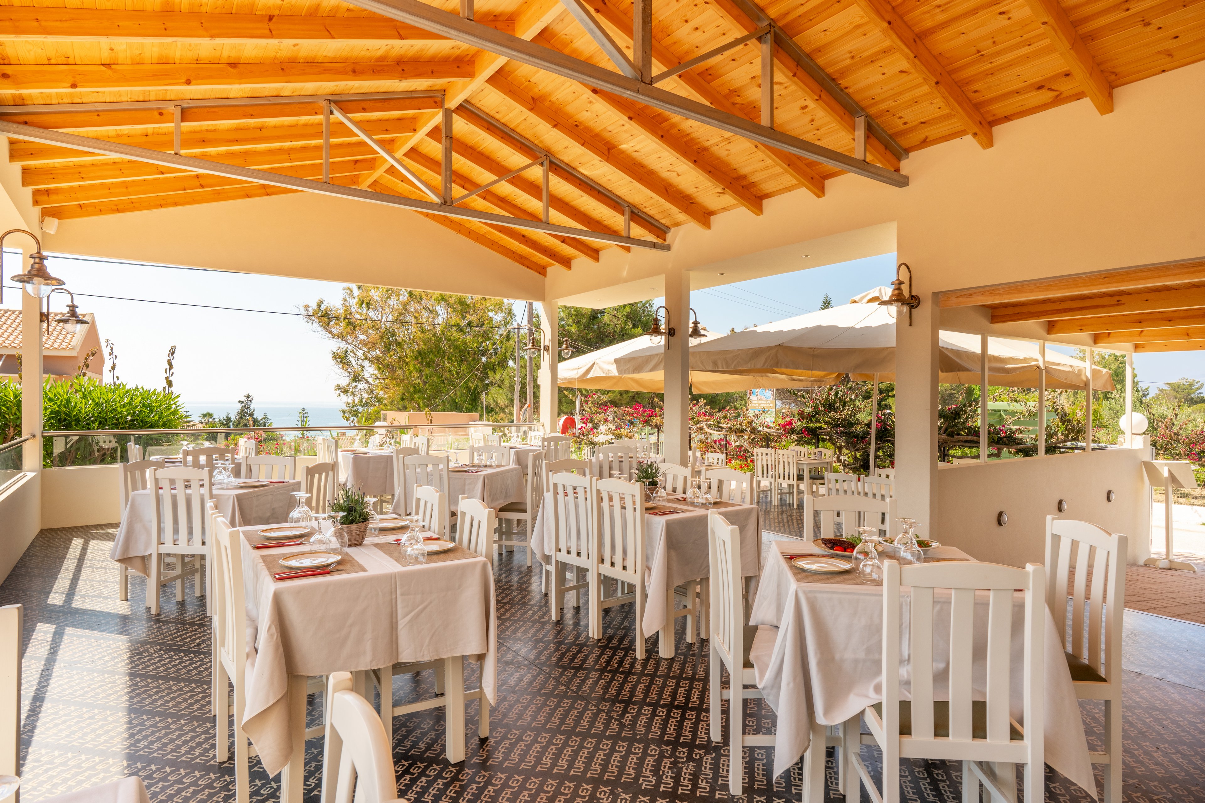 Bright outdoor restaurant terrace with neatly arranged white tables and chairs under a wooden roof. Tables are set with plates, glasses, and small decorative centerpieces, ready for guests. Large umbrellas and lush greenery surround the seating area, with a glimpse of the sea in the background. The warm sunlight and natural materials create a relaxed, inviting Mediterranean atmosphere.