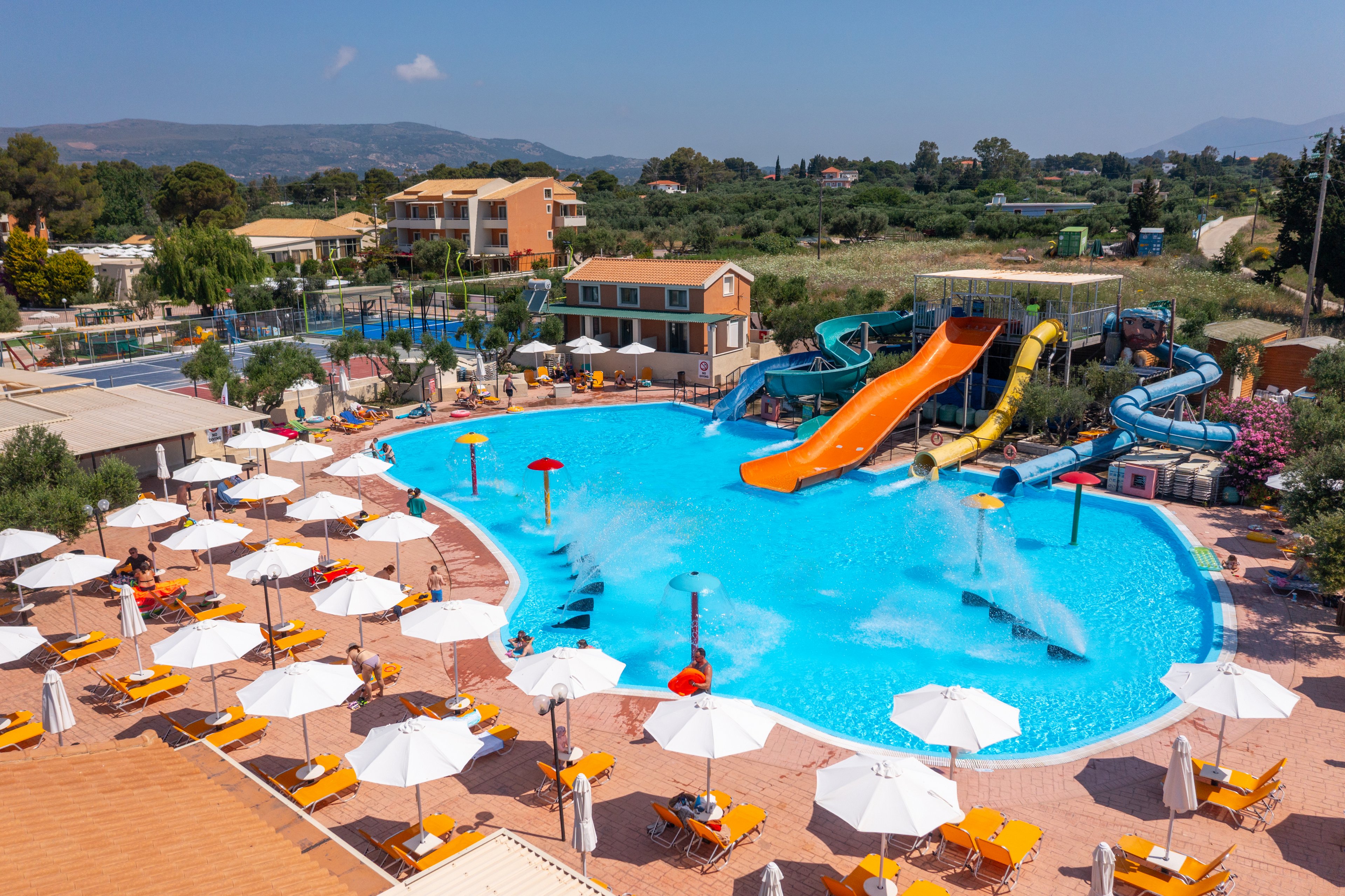 A large outdoor swimming pool is surrounded by rows of white umbrellas and sun loungers at a holiday resort. Several bright orange and blue waterslides curve into the pool, adding a playful focal point. People relax on the deck while water fountains spray into the clear blue pool. In the background, low-rise buildings and green landscape extend toward distant hills under a clear sky.