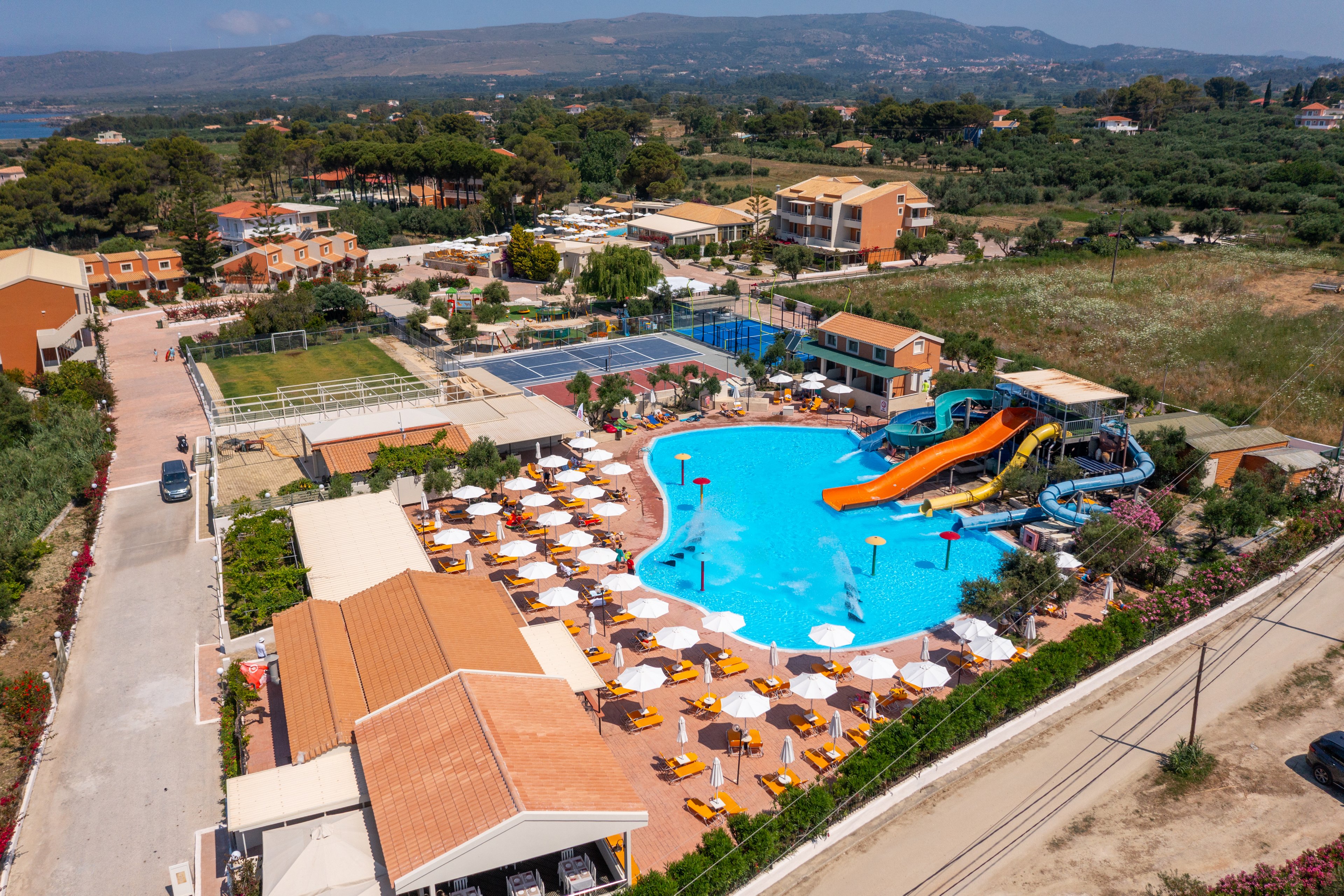 A large resort swimming pool with bright blue water is seen from above, surrounded by rows of sun loungers and umbrellas. Multiple colorful waterslides curve into the pool, creating a lively focal point. The complex includes nearby tennis courts, additional buildings, and landscaped paths, all set within a green countryside. Hills and distant mountains frame the background under a clear sky, suggesting a sunny vacation destination.