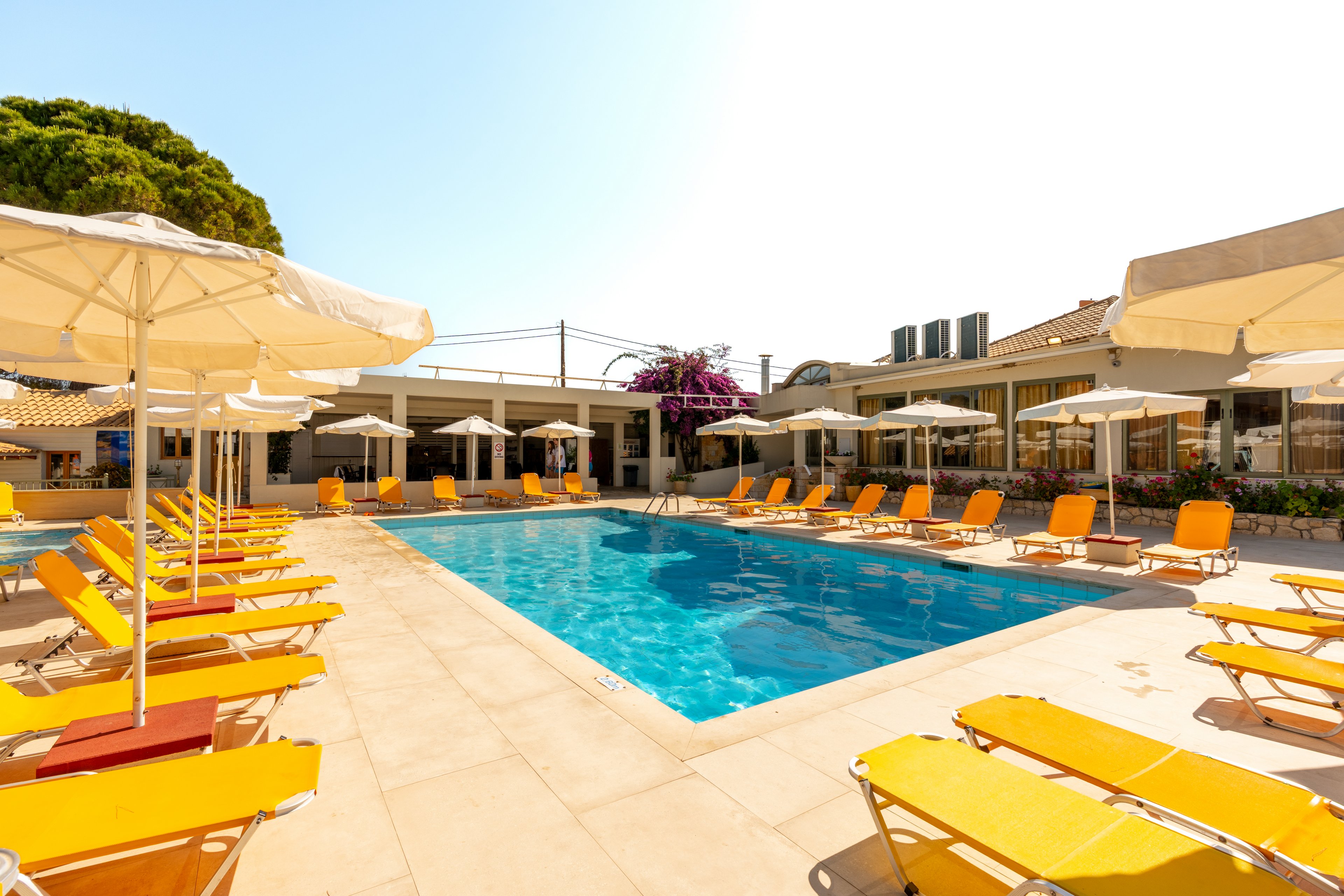 An outdoor swimming pool is surrounded by bright yellow sun loungers and large white umbrellas at a resort. The scene is set on a spacious tiled deck under clear, sunny skies. Low-rise buildings and flowering plants frame the pool area, with a few taller city buildings visible in the background. The overall atmosphere is relaxed and inviting, suggesting a vacation or leisure setting.