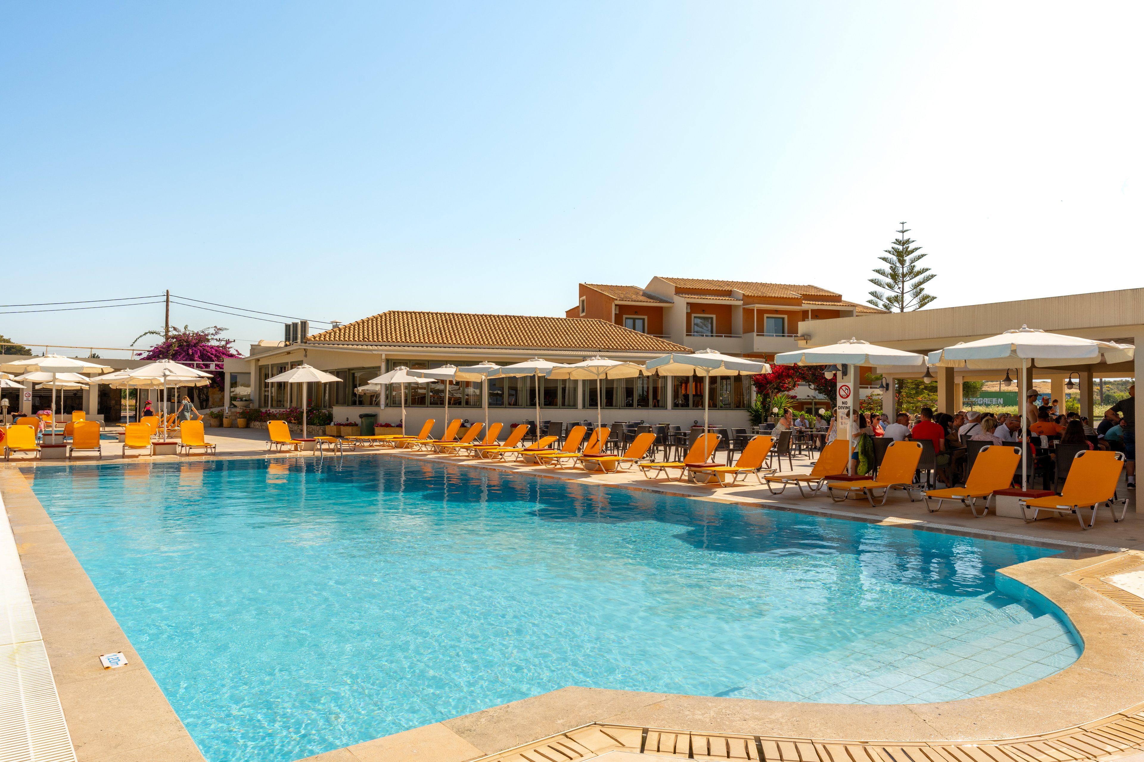 An outdoor swimming pool is surrounded by rows of orange sun loungers and white umbrellas at a resort. Guests relax in the shaded seating area near the pool, enjoying the clear, sunny weather. Low-rise buildings with tiled roofs and flowering plants frame the background, creating a vacation atmosphere. The bright blue water and cloudless sky emphasize a warm, inviting holiday setting.