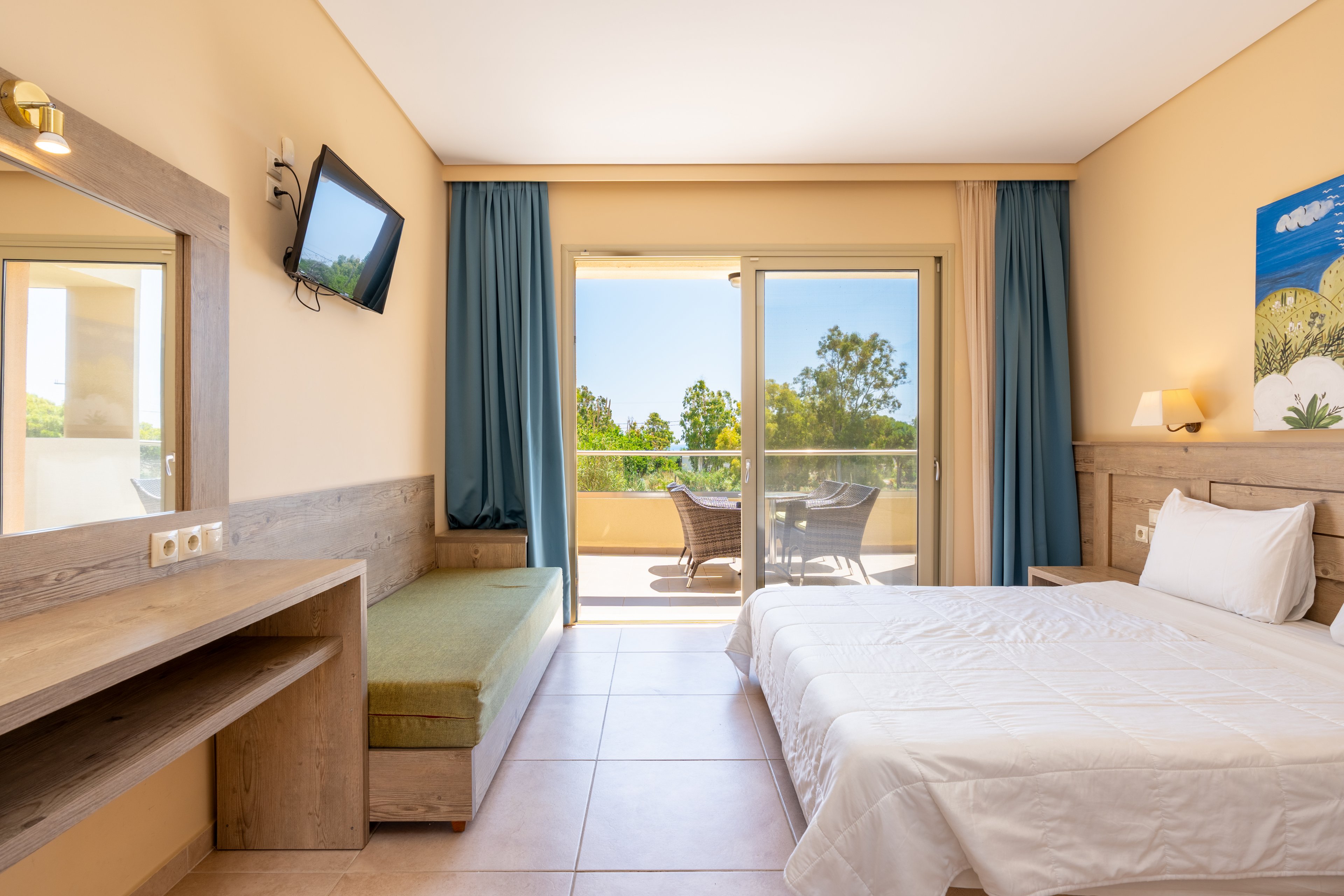 A sunlit hotel bedroom features a neatly made double bed with white linens and a wooden headboard. A desk with a large mirror and a green cushioned bench lines one wall, while a flat-screen TV is mounted above. Floor-to-ceiling glass doors open onto a balcony with outdoor chairs and a table, overlooking lush green trees under a clear blue sky. Soft beige walls and blue curtains create a calm, inviting atmosphere.