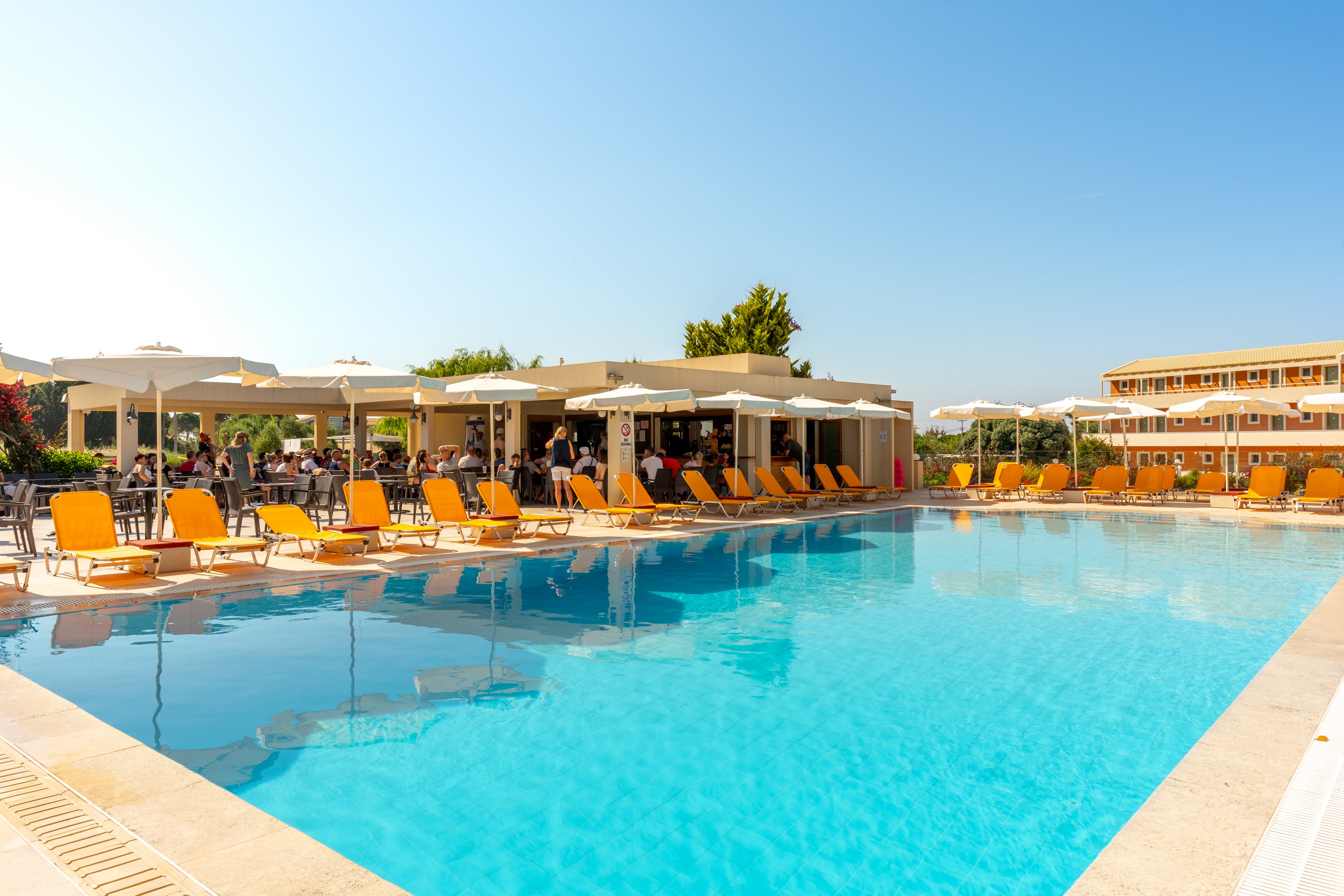 Large outdoor swimming pool at a resort is surrounded by bright yellow sun loungers and white umbrellas. Guests relax near the poolside bar and seating area under clear blue skies. Modern low-rise buildings frame the background, creating a vacation atmosphere. The scene conveys a warm, sunny day ideal for leisure and relaxation.